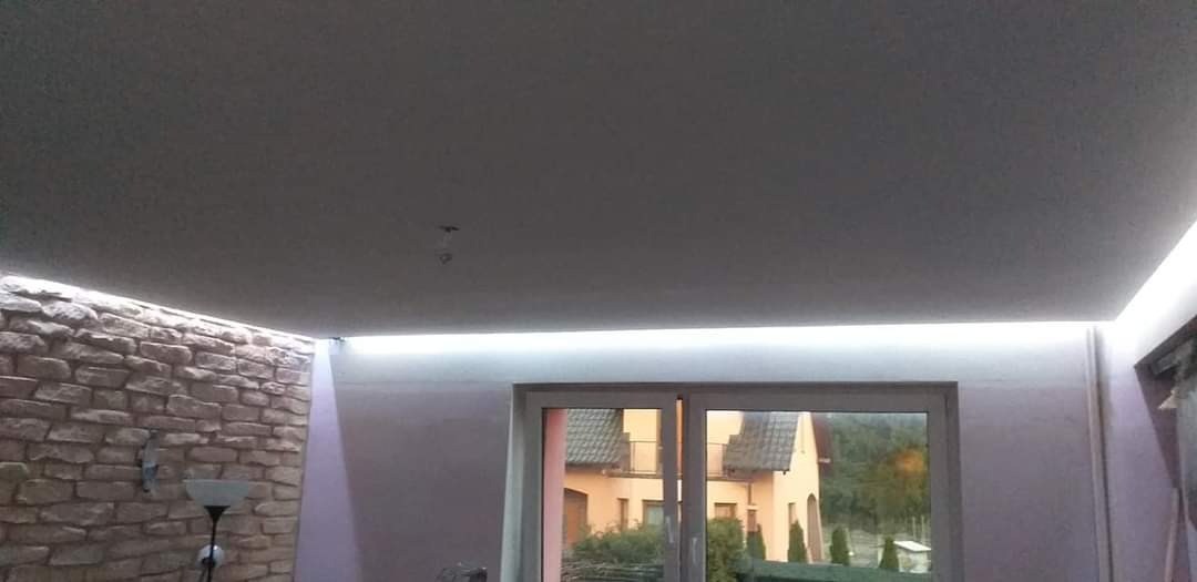 Wykończony balkon z oświetleniem LED pod sufitem, ściana wyłożona kamieniem dekoracyjnym, widok na dom przez okno balkonowe.