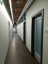 Korytarz biurowy z białymi ścianami, ciemną podłogą, oświetleniem LED pod sufitem i drzwiami z ciemnymi ościeżnicami. Widoczne fragmenty instalacji pod sufitem.