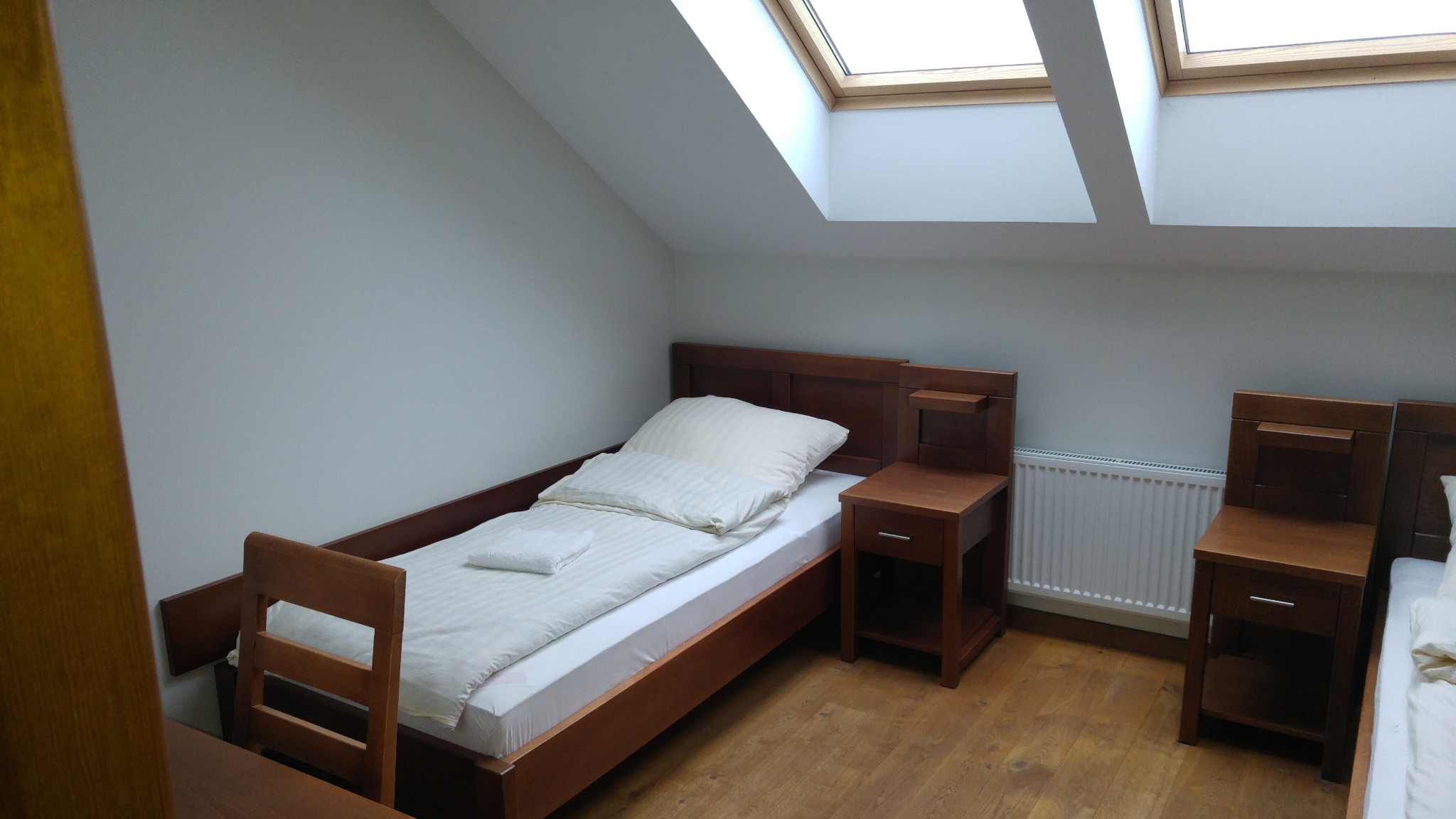 Pokój na poddaszu z dwoma pojedynczymi łóżkami z drewnianymi ramami i szafkami nocnymi, oświetlony przez okna dachowe.