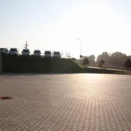 Rozległy parking wyłożony szarą kostką brukową w jodełkę, w tle rząd zaparkowanych białych busów na wzgórzu porośniętym trawą, w oddali drzewa i słup oświetleniowy.