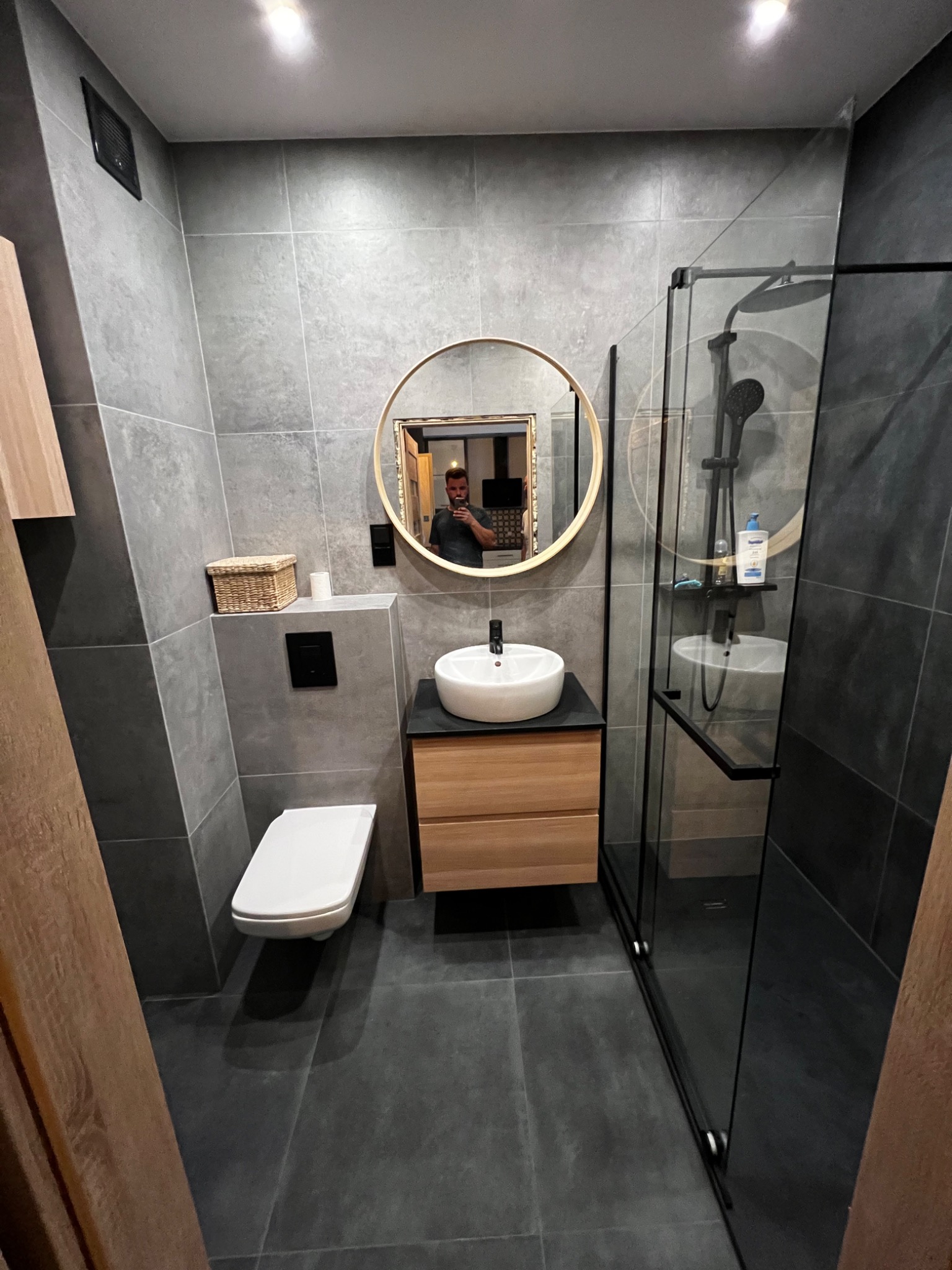 Nowoczesna łazienka w szarościach z okrągłym lustrem, kabiną prysznicową typu walk-in i wiszącą toaletą oraz szafką z umywalką z drewnianymi frontami.