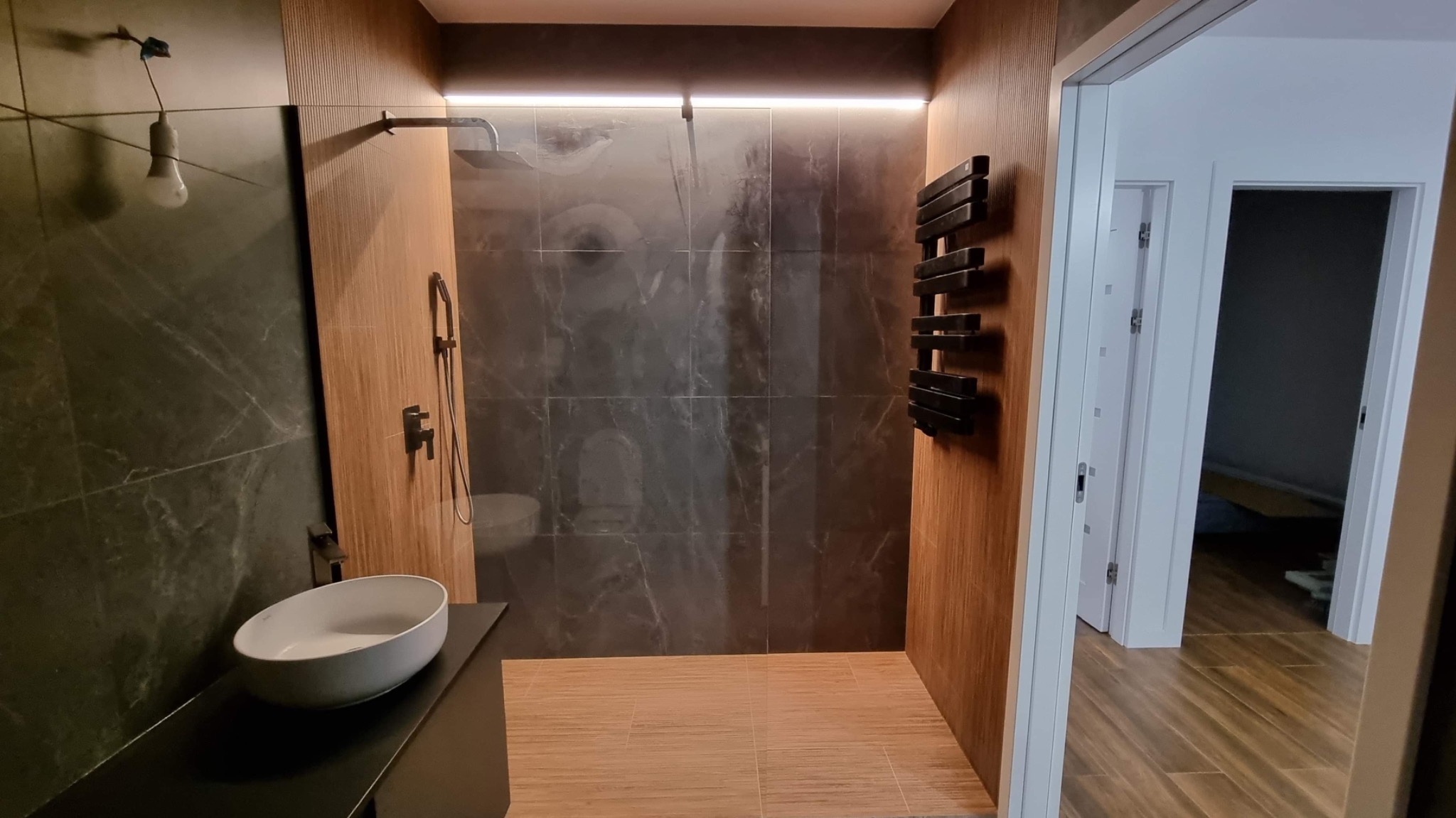 Nowoczesna łazienka z marmurowymi płytkami pod prysznicem, drewnianymi panelami i minimalistyczną umywalką nablatową, oświetlona listwą LED.
