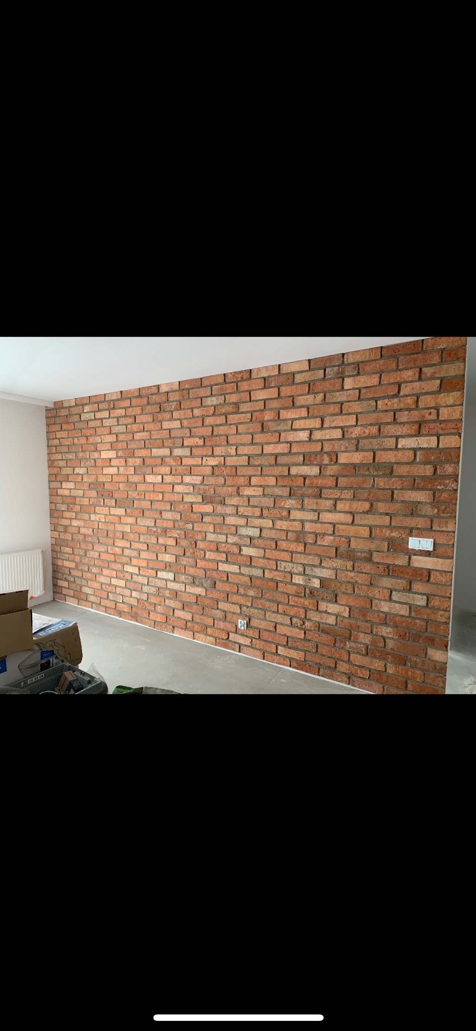 Ściana wewnętrzna wykończona dekoracyjną czerwoną cegłą, widoczne gniazdka elektryczne i fragment grzejnika, podłoga w trakcie przygotowania.