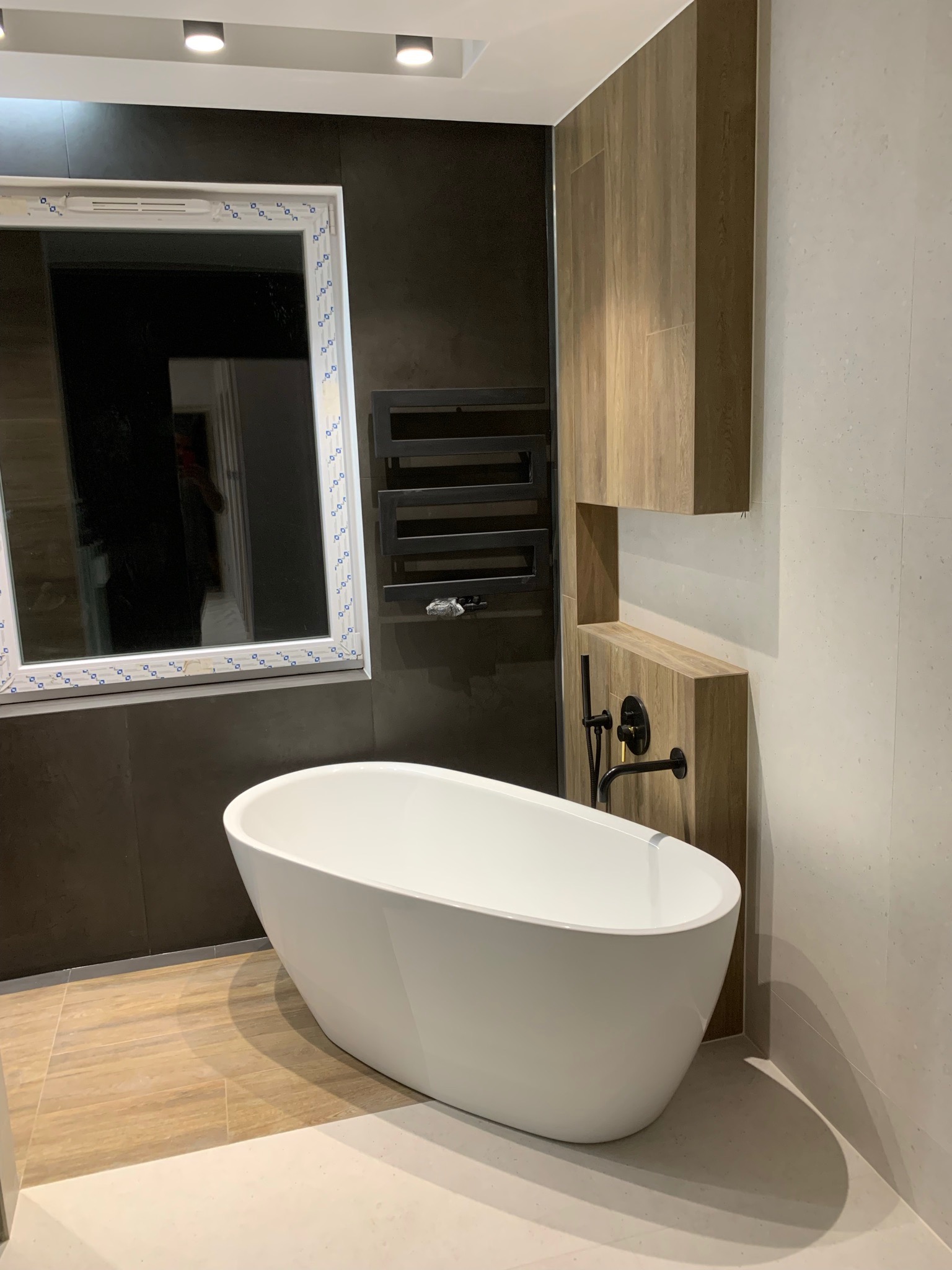 Nowoczesna łazienka z wolnostojącą białą wanną, czarnym grzejnikiem drabinkowym i drewnopodobnymi elementami zabudowy, widok na okno z białymi ramami.
