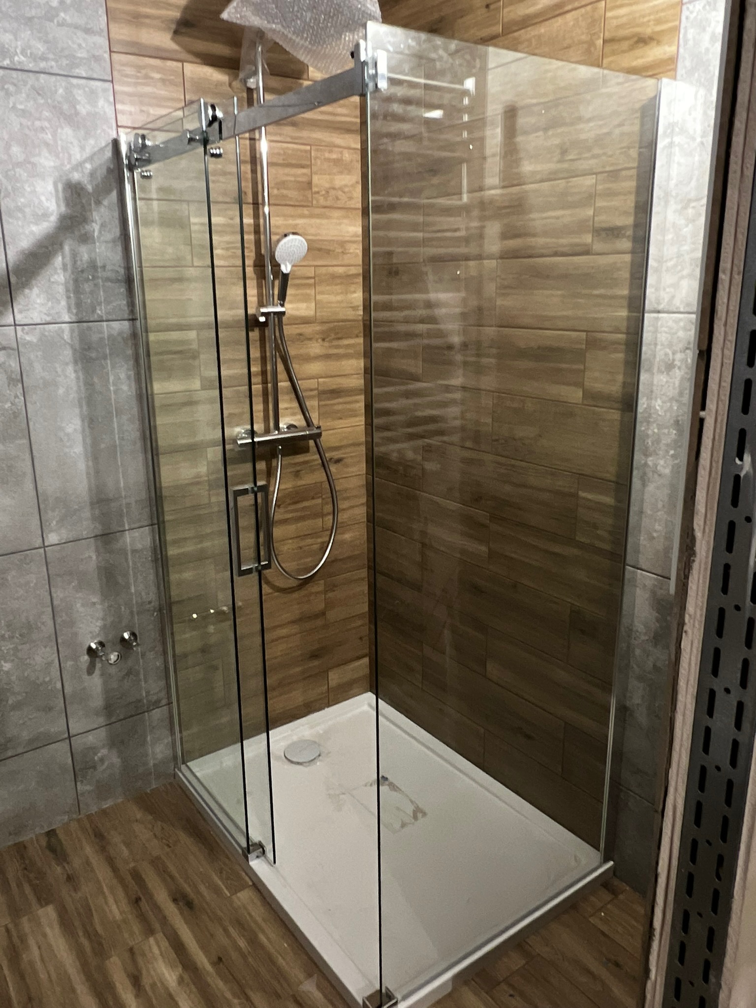Nowoczesna kabina prysznicowa z przesuwnymi drzwiami szklanymi, zamontowana na białym brodziku, z widoczną armaturą w kolorze chromu, na tle płytek imitujących drewno i szarego kamienia.