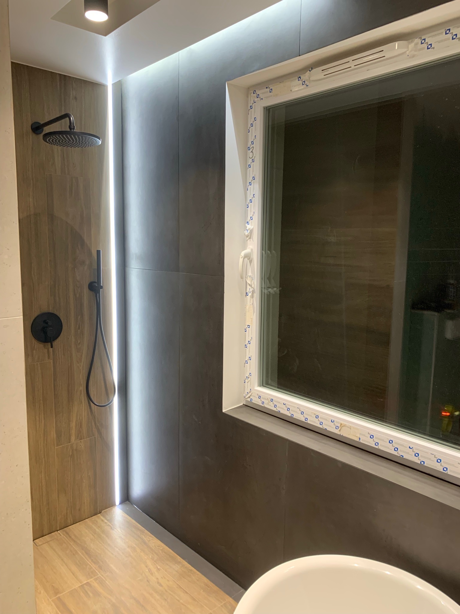 Nowoczesna łazienka z czarną armaturą prysznicową, oświetleniem LED i oknem z folią ochronną na ramie, ukazująca wykończone wnętrze.