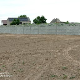 Profi ViOl Company Viktor Savchuk - Długi, szary mur z prefabrykowanych elementów betonowych, z dekoracyjnym ażurowym wykończeniem górnej krawędzi, oddziela pole od zabudowań mieszkalnych w tle.