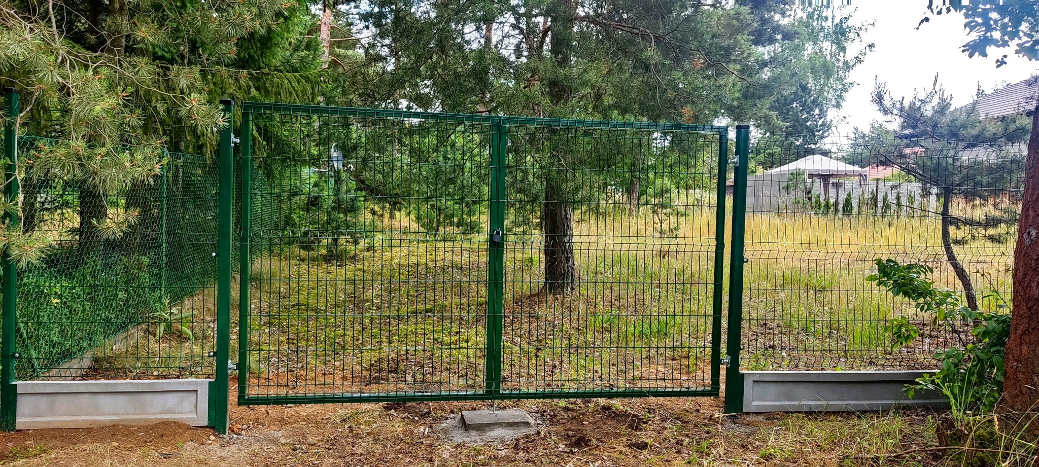 Zielona, dwuskrzydłowa brama z siatki panelowej, z betonową podmurówką, zamontowana na tle leśnej działki z widoczną altaną.