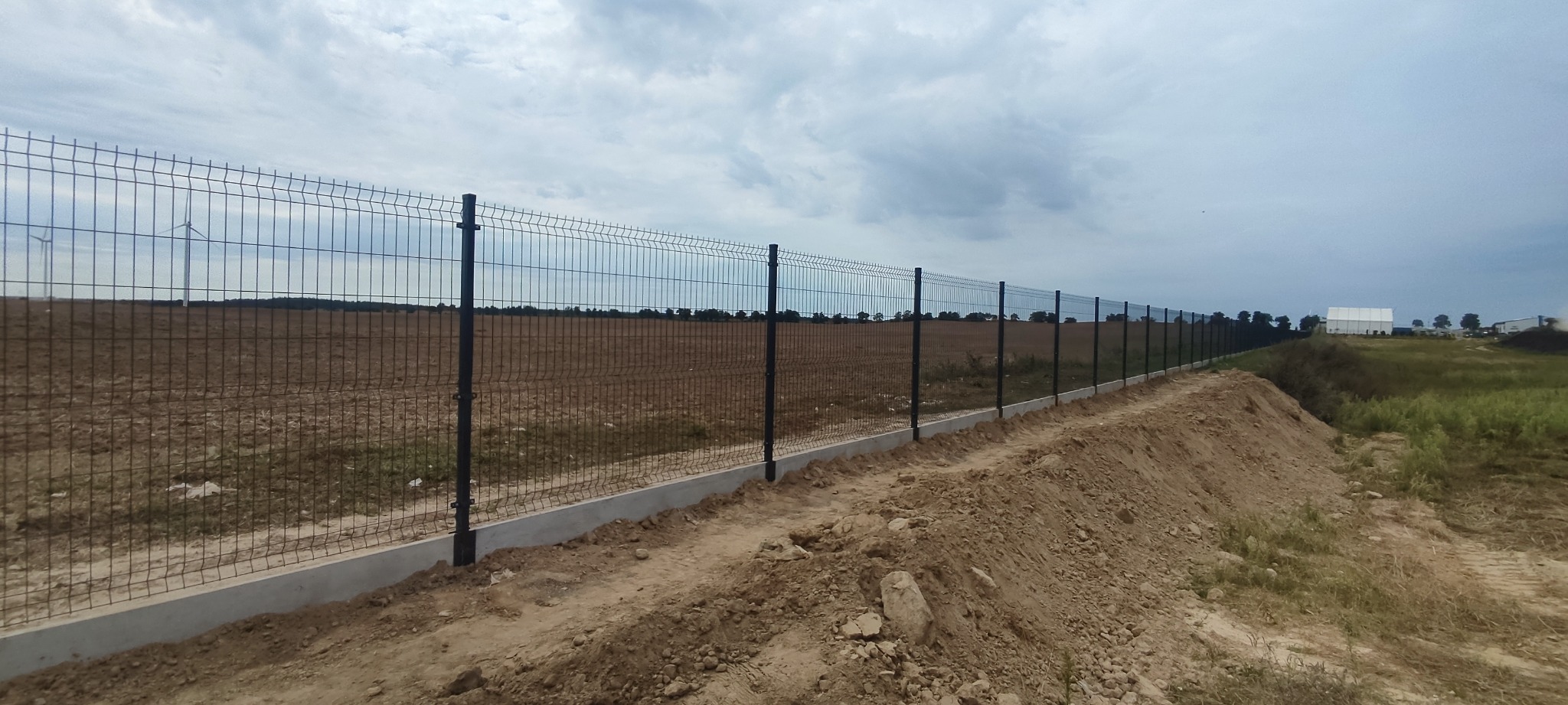 Długi, ciemny system ogradzający z siatki panelowej na betonowej podmurówce, biegnący wzdłuż pola w pochmurny dzień, z turbinami wiatrowymi widocznymi w oddali.