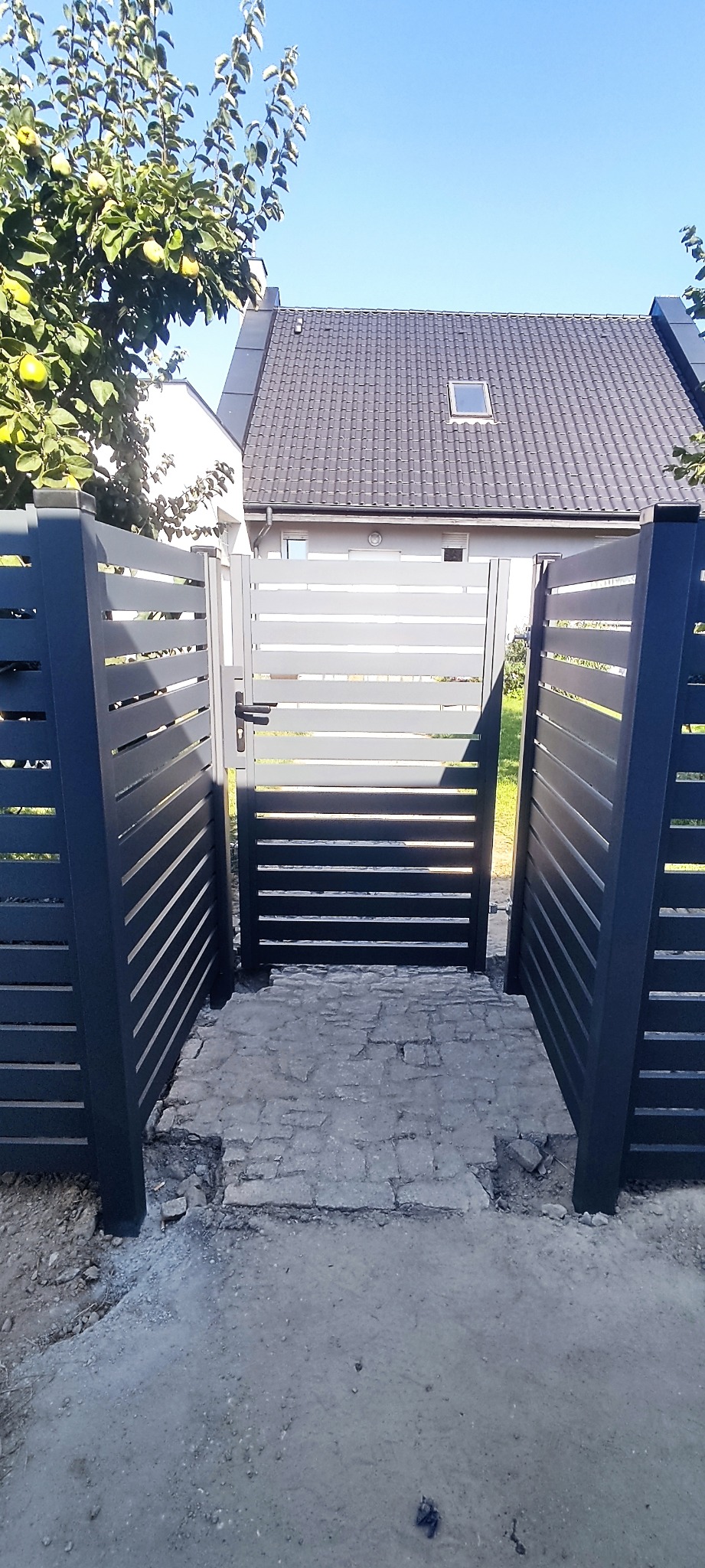 Nowoczesna brama z poziomych profili, prowadząca do domu z szarą dachówką, widok z zewnątrz na częściowo ułożoną kostkę brukową.
