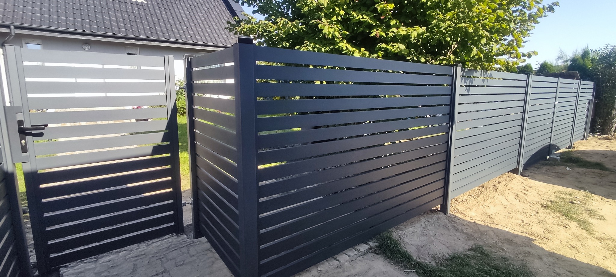 Nowoczesne, poziome ogrodzenie panelowe w kolorze grafitowym z furtką, zrealizowane w Przeźmierowie. Widoczne elementy betonowej podmurówki. Tło stanowi zielona roślinność i fragment budynku.