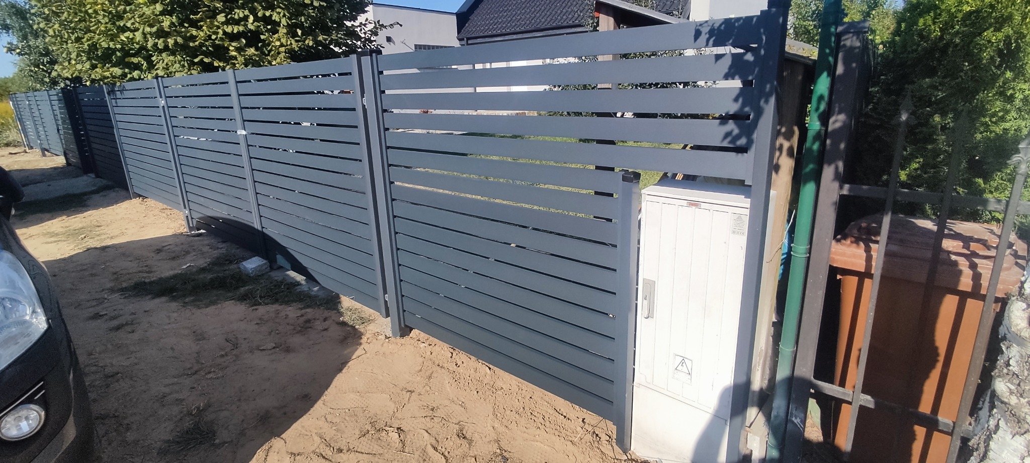 Nowoczesne, poziome, szare ogrodzenie z metalowych paneli, widok z boku, z widoczną skrzynką elektryczną i fragmentem samochodu w tle.