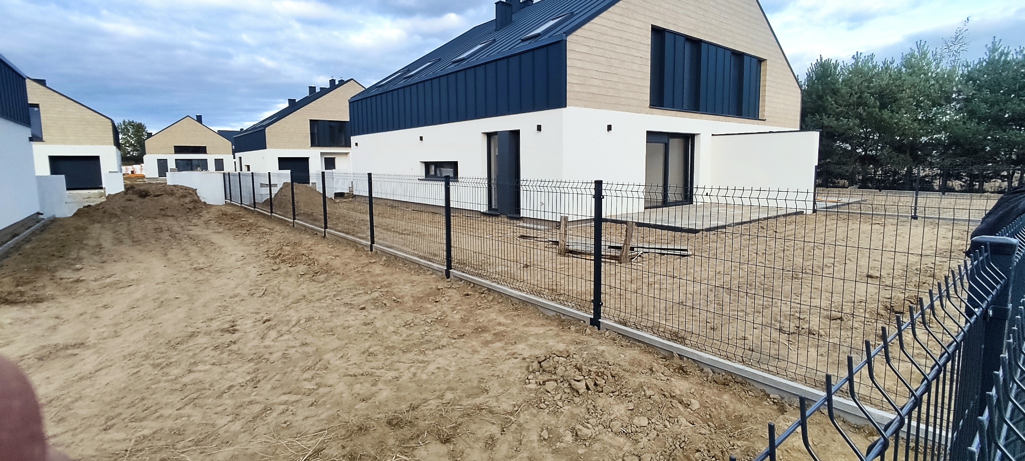 Nowoczesne domy jednorodzinne z czarnymi dachami i jasnymi elewacjami, oddzielone od siebie czarną siatką na betonowej podmurówce, teren wokół w trakcie zagospodarowywania.