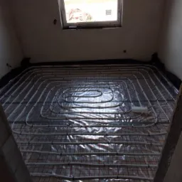 Białe rury ogrzewania podłogowego ułożone na folii z czerwoną siatką w jasnym pomieszczeniu z oknem dachowym. Widok z góry na instalację przed zalaniem wylewką.