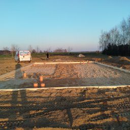 Usługi hydrauliczne Paweł Wilczyński - Przygotowanie terenu pod budowę: wytyczone fundamenty z deskowaniem, rury kanalizacyjne, piasek. W tle widać busa i sylwetkę osoby.