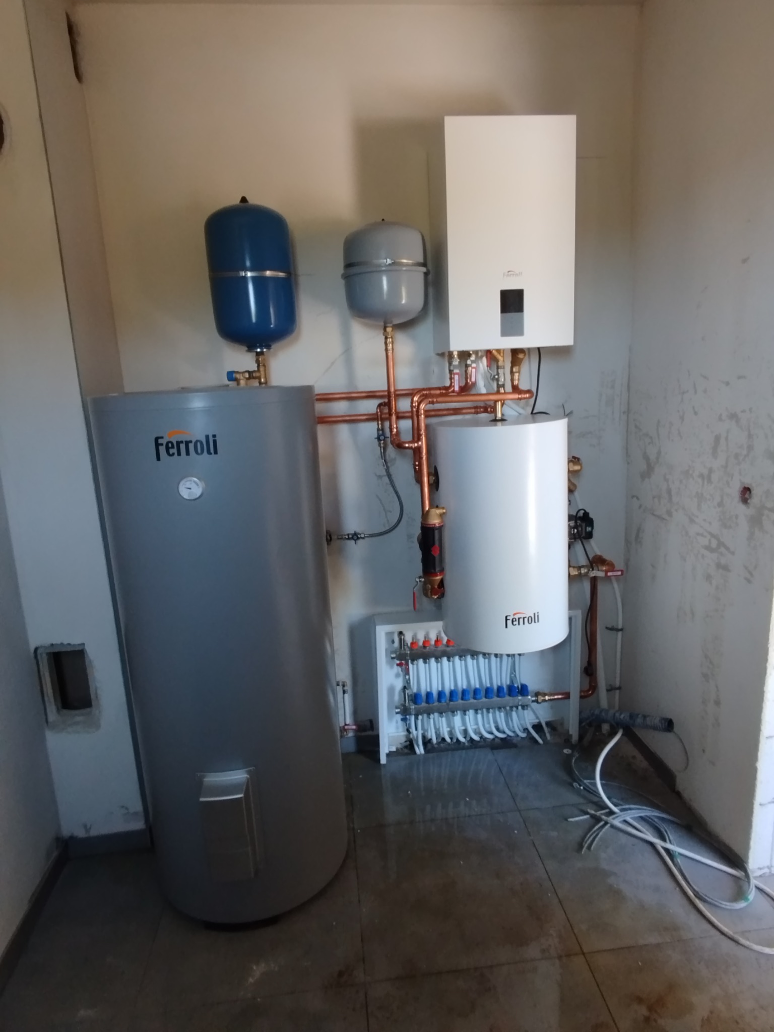 Kompletna instalacja grzewcza Ferroli: zasobnik, rozdzielacz i piec, z miedzianymi rurami. Widoczne elementy montażowe i podłączenia. Układ w surowym pomieszczeniu.