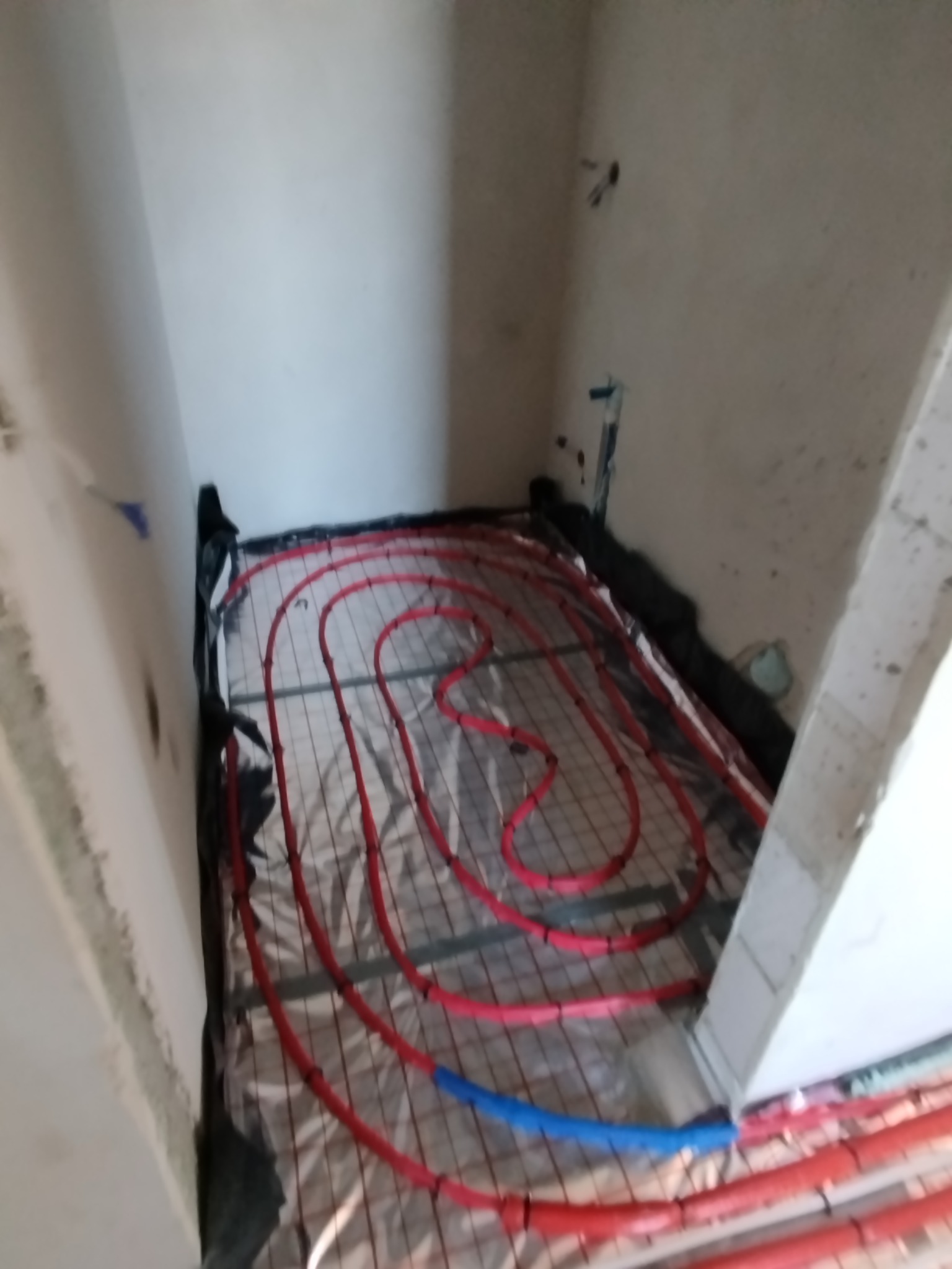 Czerwone rury ogrzewania podłogowego ułożone na srebrnej macie w surowym pomieszczeniu, widok z góry pod kątem, z elementami nieotynkowanych ścian.