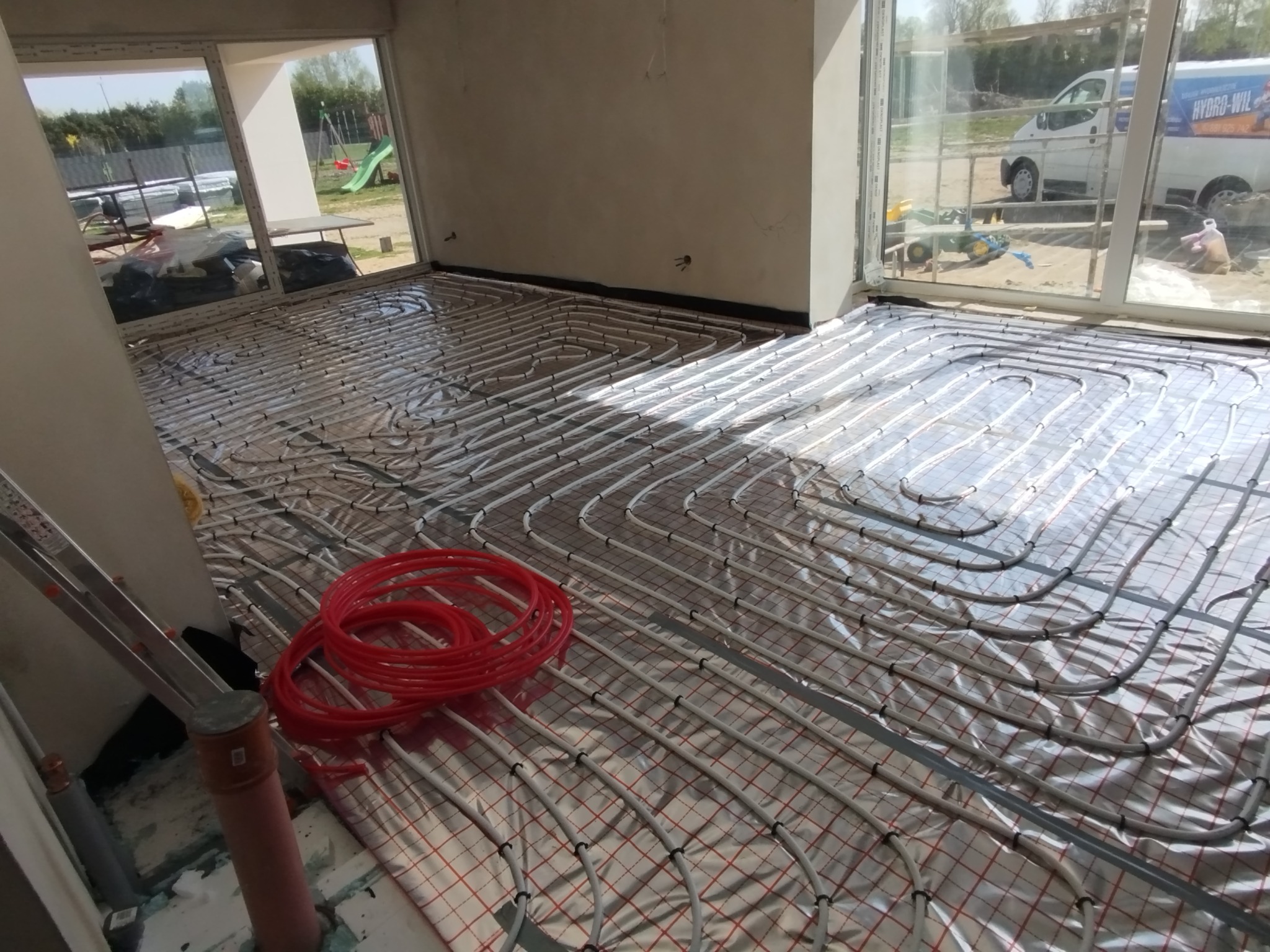 Rozłożona instalacja ogrzewania podłogowego na folii izolacyjnej z widocznymi czerwonymi rurami. W tle widoczne okno i teren budowy.