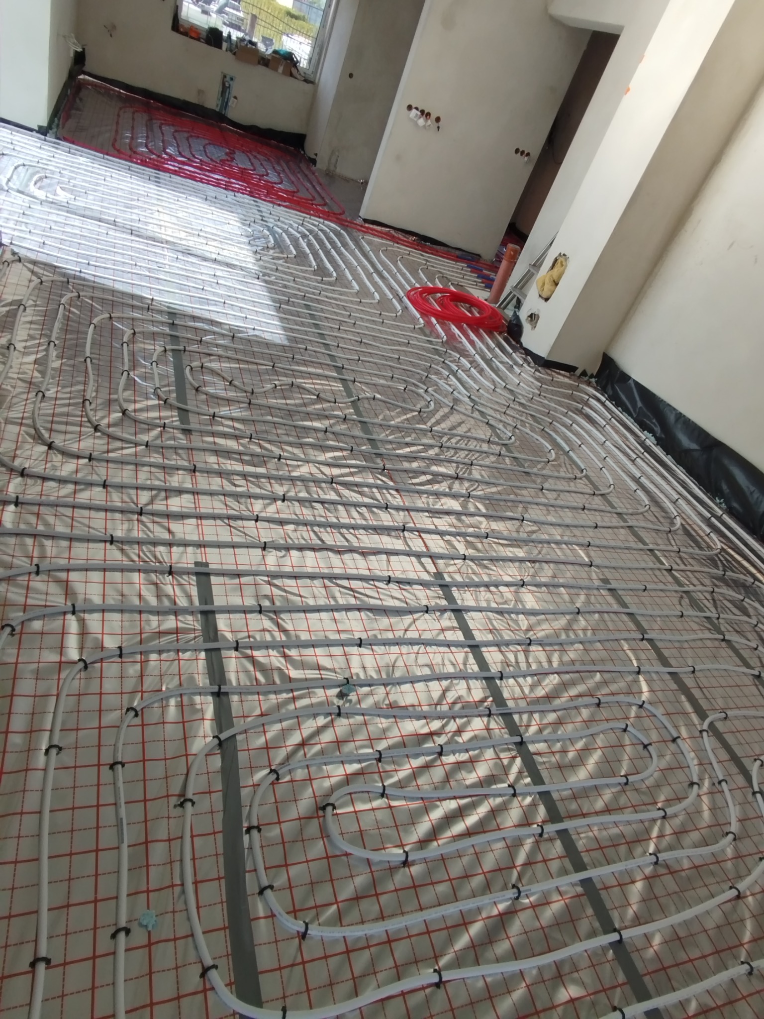 Białe rury ogrzewania podłogowego ułożone spiralnie na folii z czerwoną siatką w surowym wnętrzu, fragment czerwonej rury. Widok z góry, ukośny kadr.