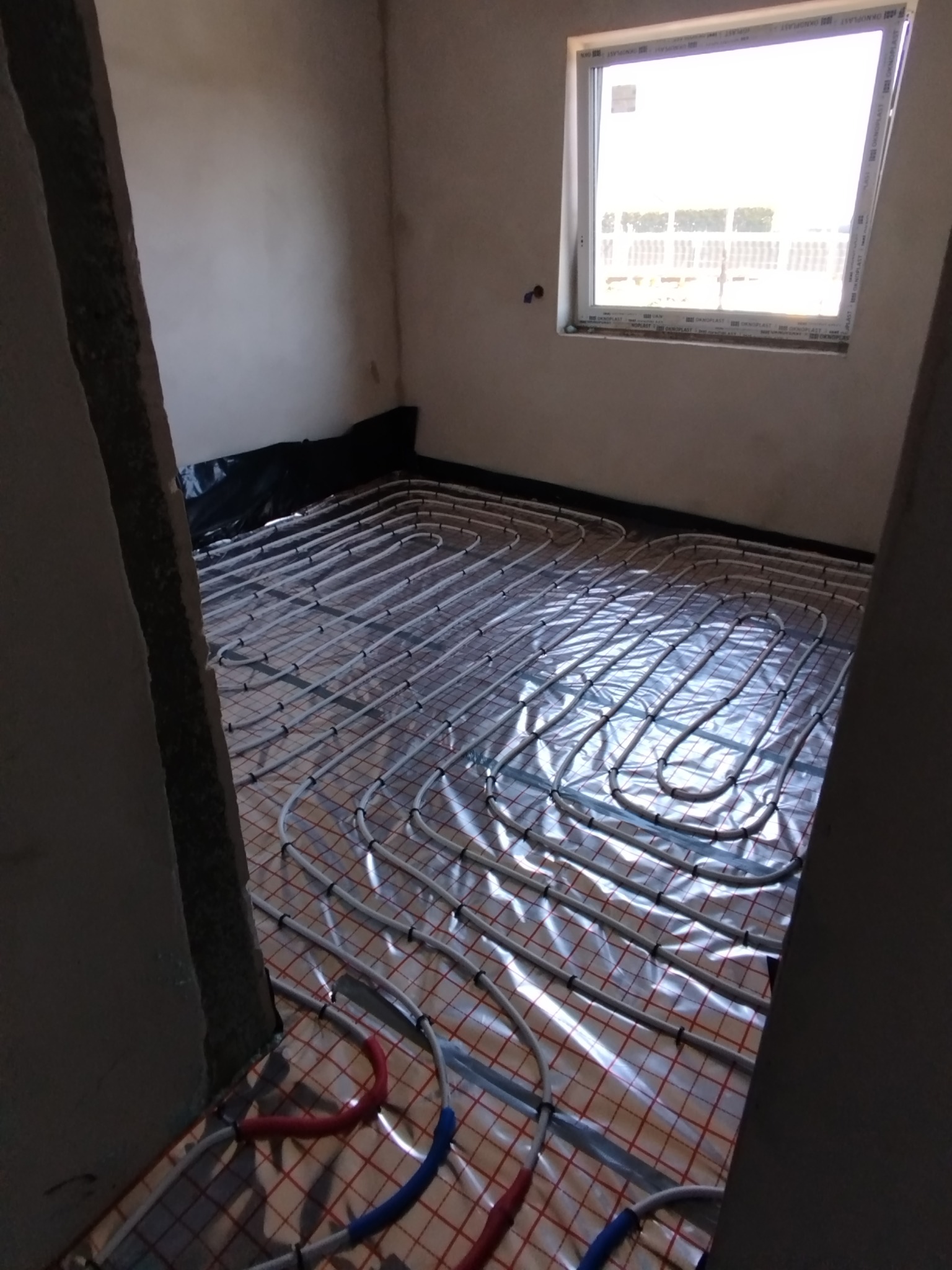Białe rury ogrzewania podłogowego rozłożone na folii w surowym pomieszczeniu z oknem, widoczne fragmenty ścian i podłogi, początkowy etap instalacji.