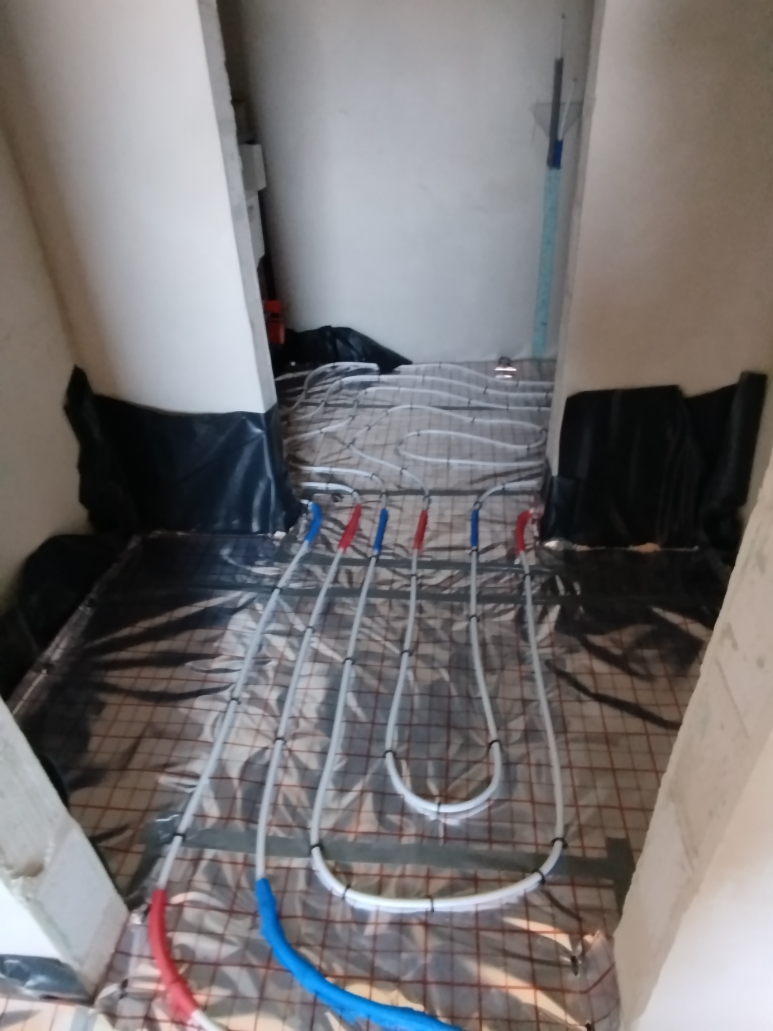 Ułożona instalacja ogrzewania podłogowego na folii z rastrem, widoczne rury z oznaczeniami kolorem czerwonym i niebieskim. Fragment ściany z poziomnicą.