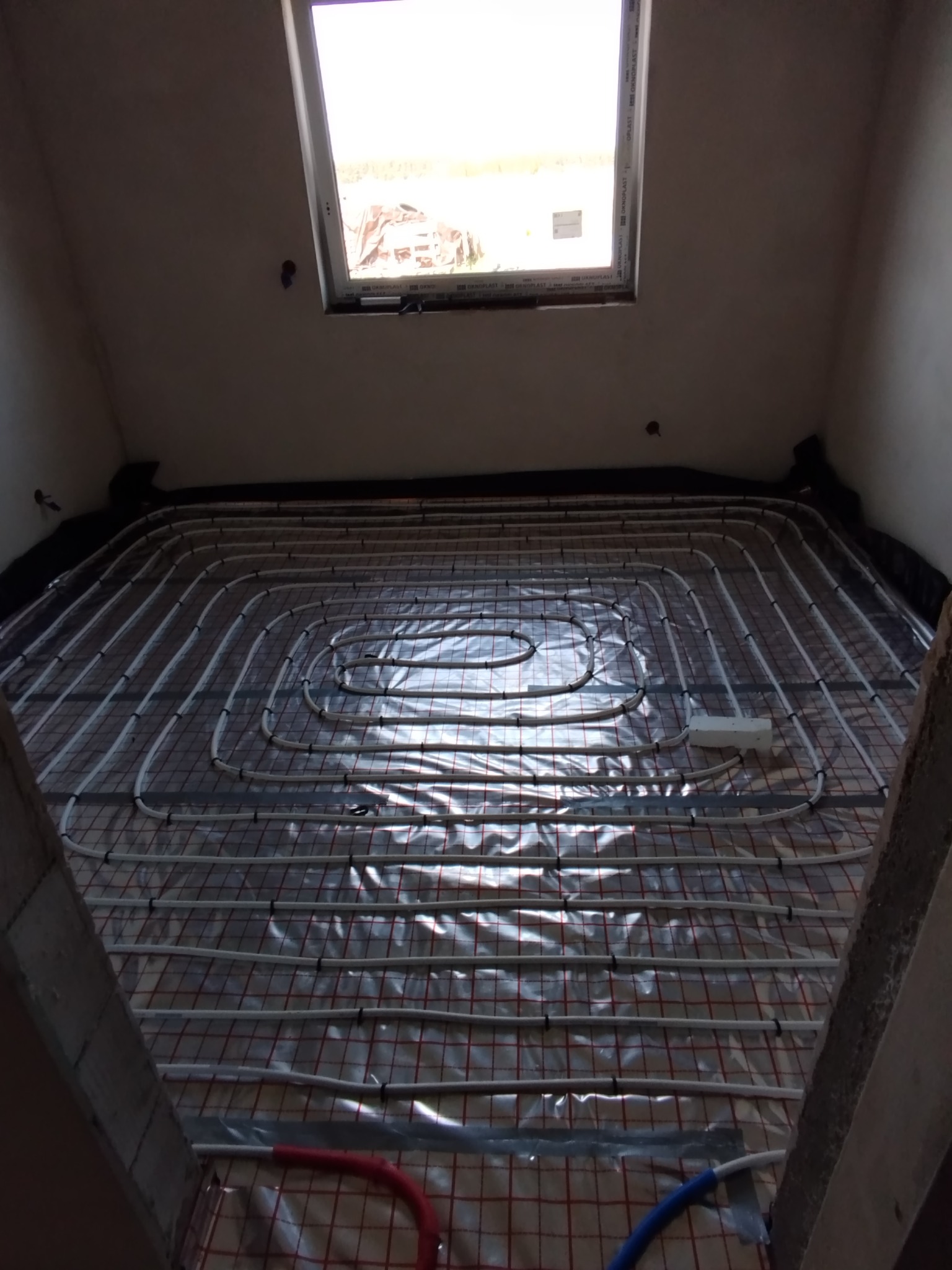 Białe rury ogrzewania podłogowego ułożone na folii z czerwoną siatką w jasnym pomieszczeniu z oknem dachowym. Widok z góry na instalację przed zalaniem wylewką.