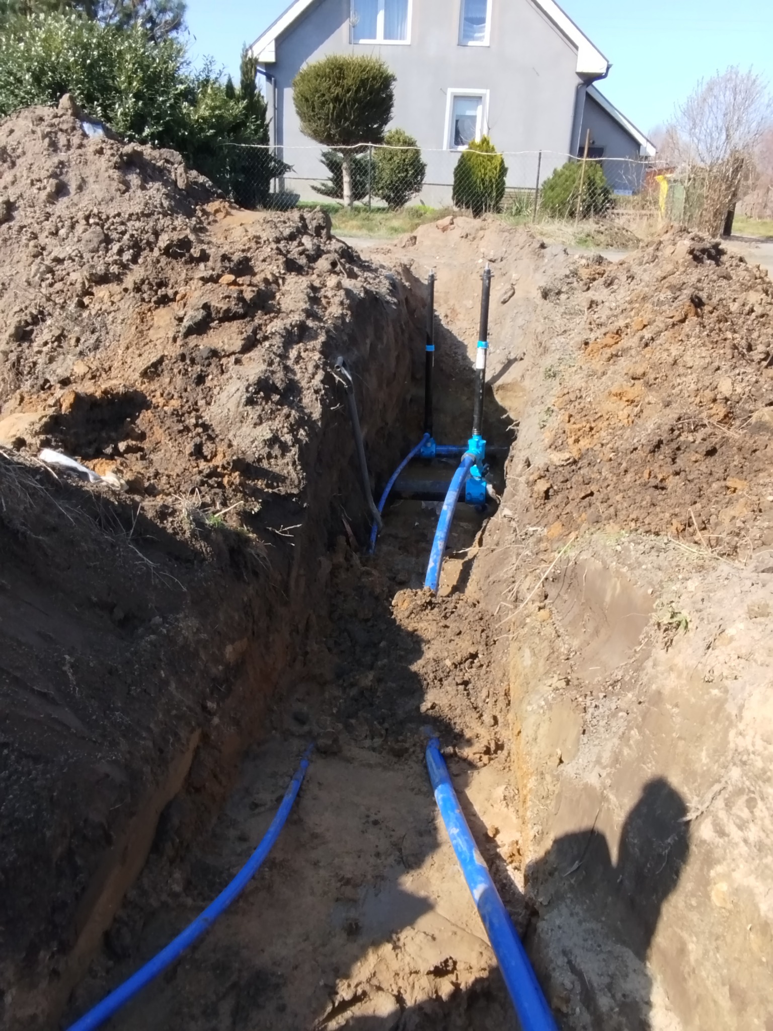 Wykop z ułożonymi niebieskimi rurami instalacji wodnej, widoczne złącza i pionowe wyprowadzenia. W tle ziemia, dom jednorodzinny i drzewa.