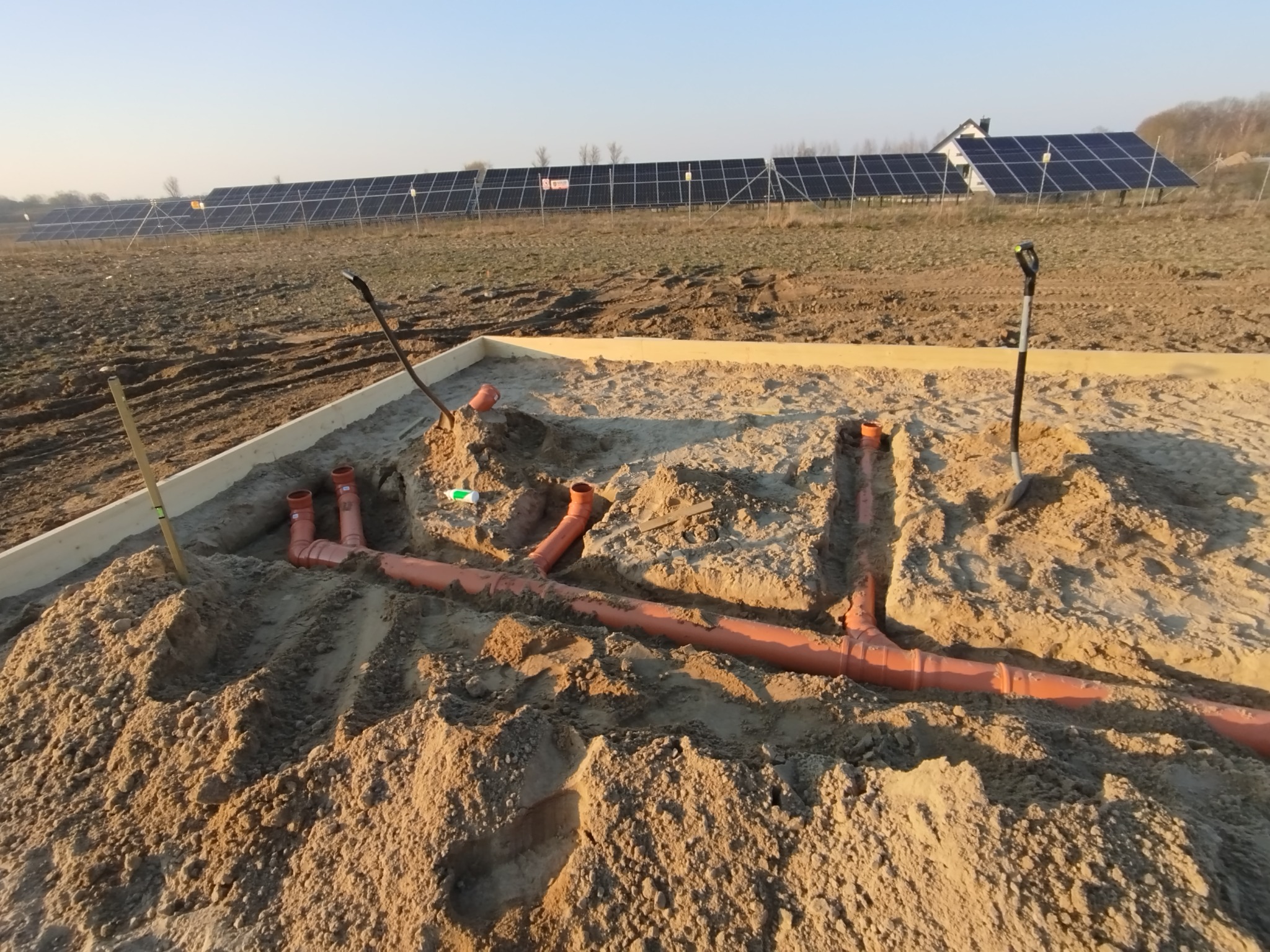 Instalacja kanalizacyjna z pomarańczowych rur w wykopie z piasku, widok na panele słoneczne w tle. Etap prac ziemnych i układania rur.