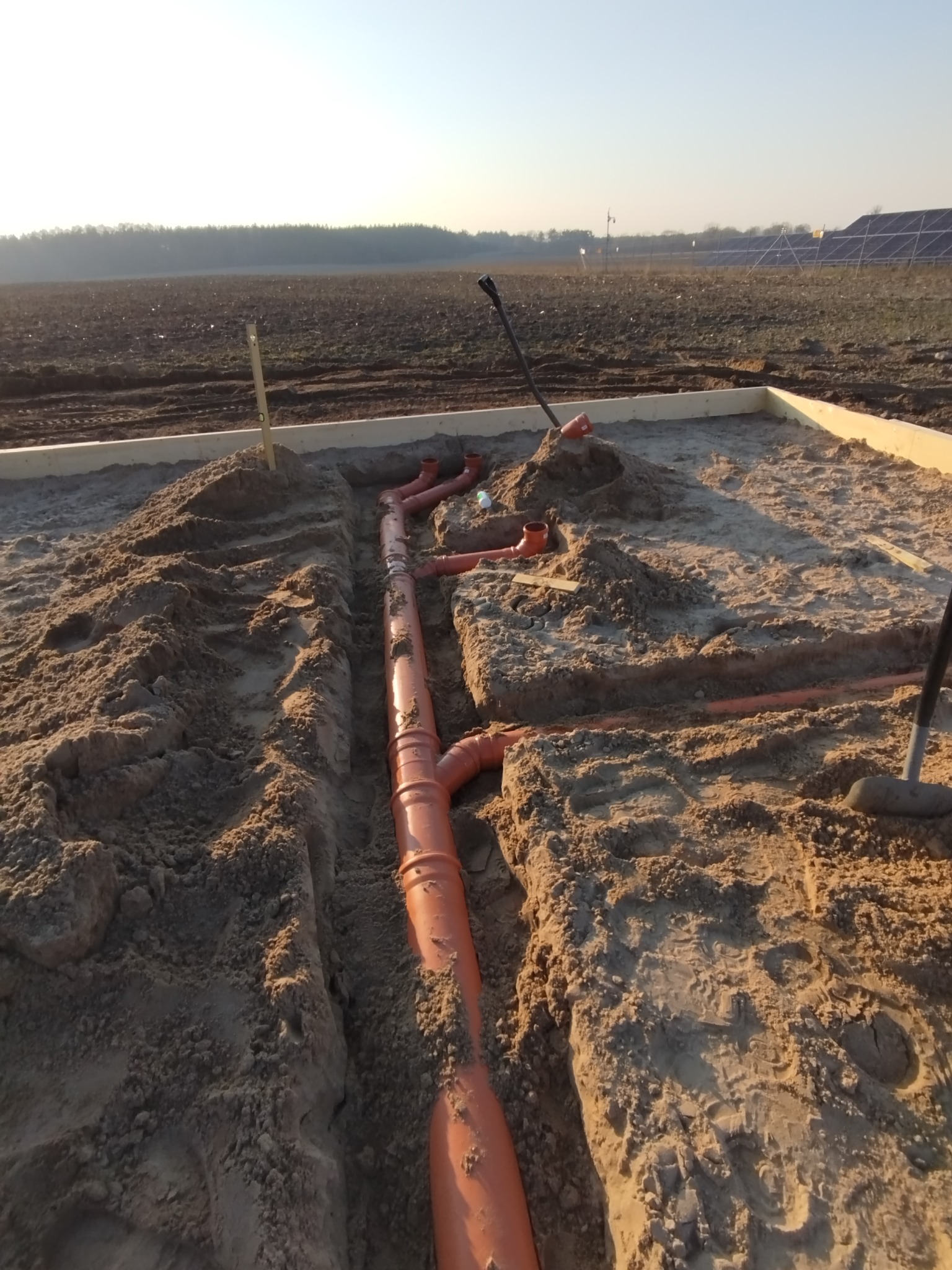 Pomarańczowe rury kanalizacyjne ułożone w wykopanym rowie, w piasku, na terenie budowy z widocznymi panelami słonecznymi w tle.