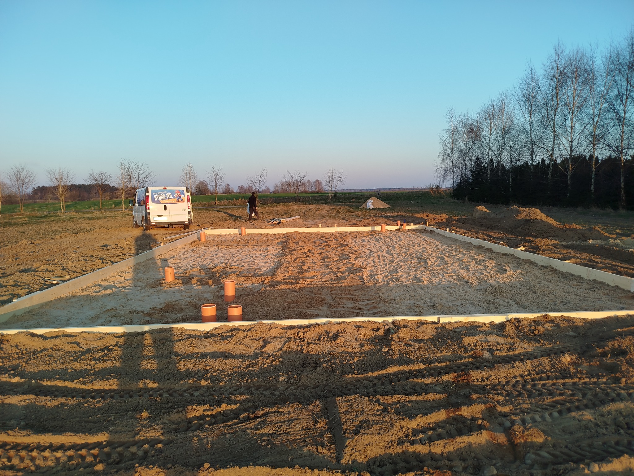 Przygotowanie terenu pod budowę: wytyczone fundamenty z deskowaniem, rury kanalizacyjne, piasek. W tle widać busa i sylwetkę osoby.