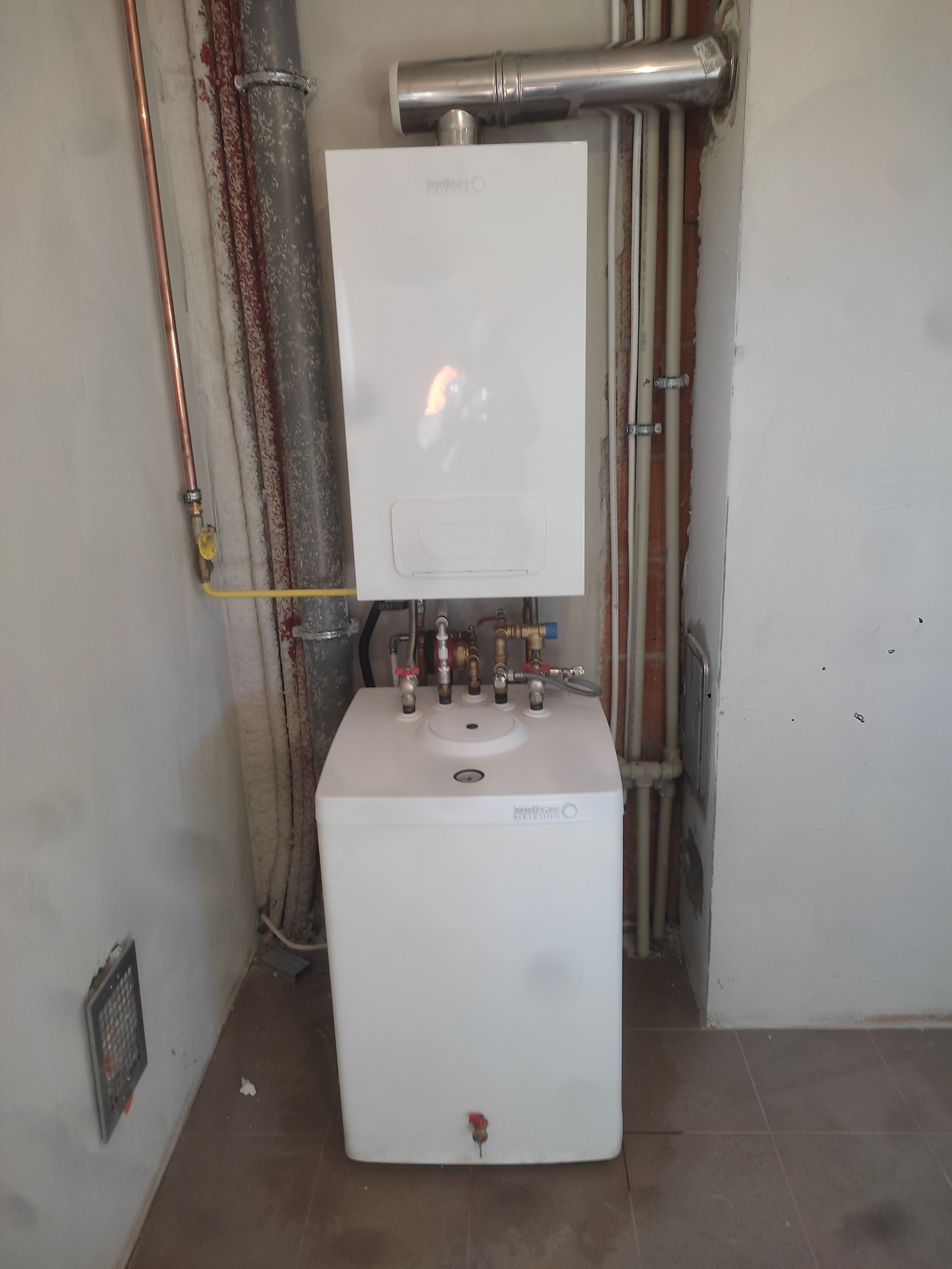 Biały, dwuczęściowy piec gazowy zamontowany na ścianie w surowym pomieszczeniu, widoczne rury miedziane i instalacyjne oraz kratka wentylacyjna w ścianie.