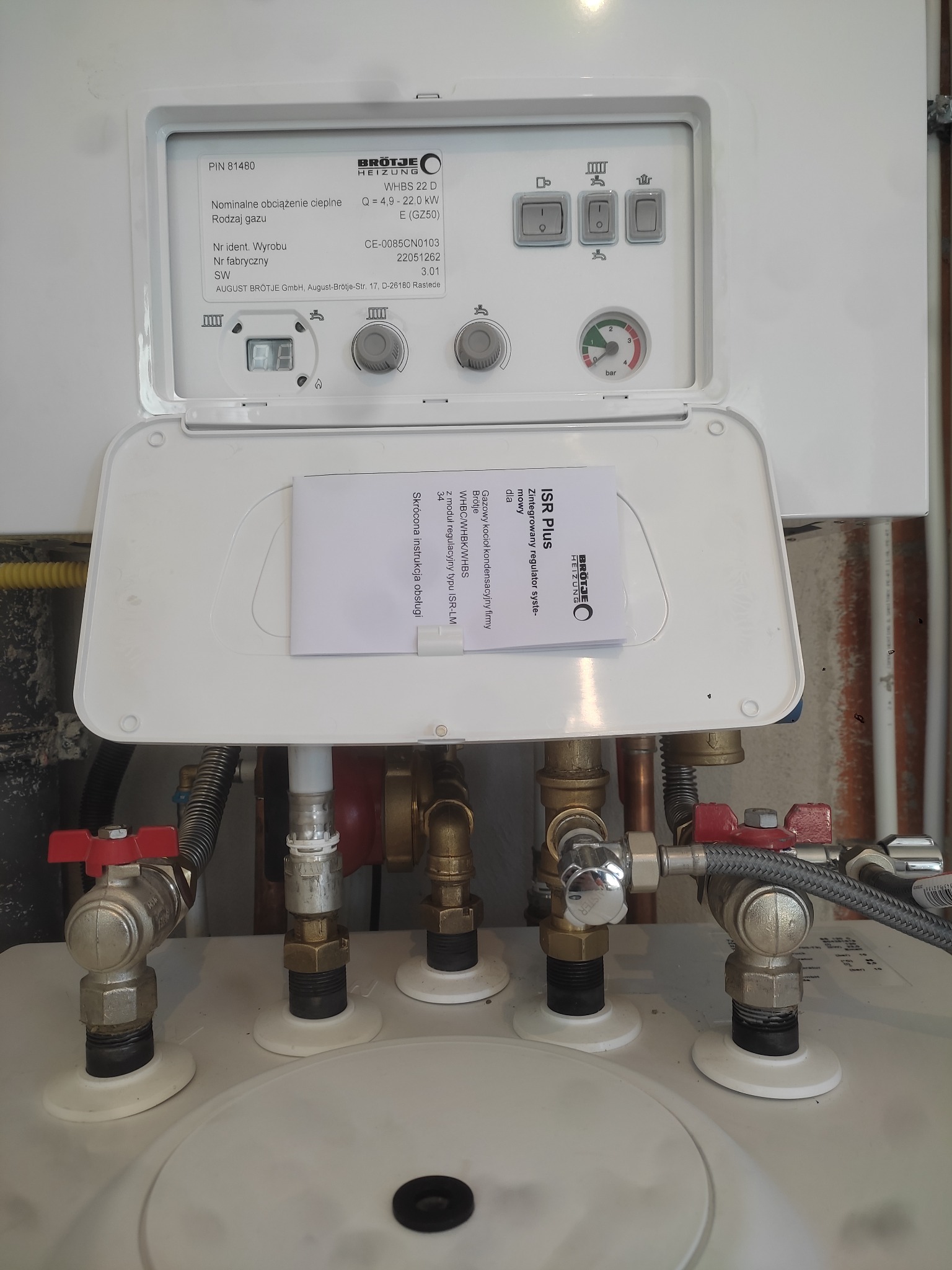 Kocioł gazowy marki Brötje z otwartą klapą panelu sterowania, widoczne przyłącza hydrauliczne z zaworami, instrukcja obsługi wewnątrz klapy.