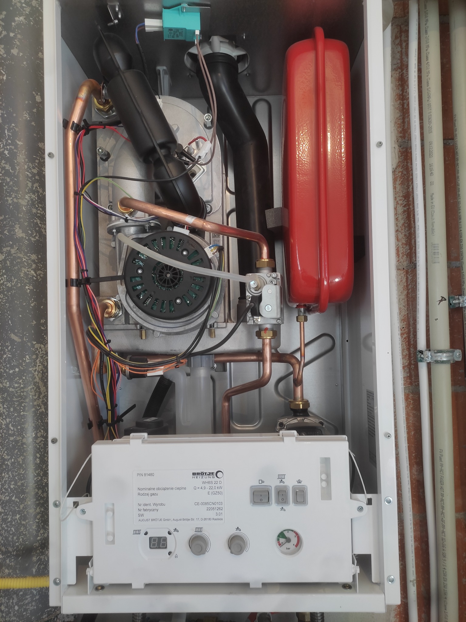 Wnętrze kotła gazowego marki Brötje WHBS 22 D z widocznymi miedzianymi rurami, czarnymi elementami instalacji i czerwonym zbiornikiem wyrównawczym, zamontowanego na ścianie z fragmentem instalacji...