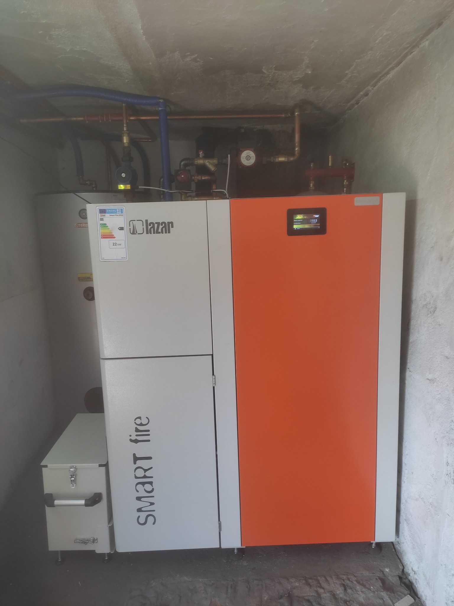 Nowoczesny piec na paliwo stałe marki Lazar Smart Fire SF22 z zasobnikiem, zainstalowany w kotłowni o surowych ścianach i widoczną instalacją hydrauliczną.