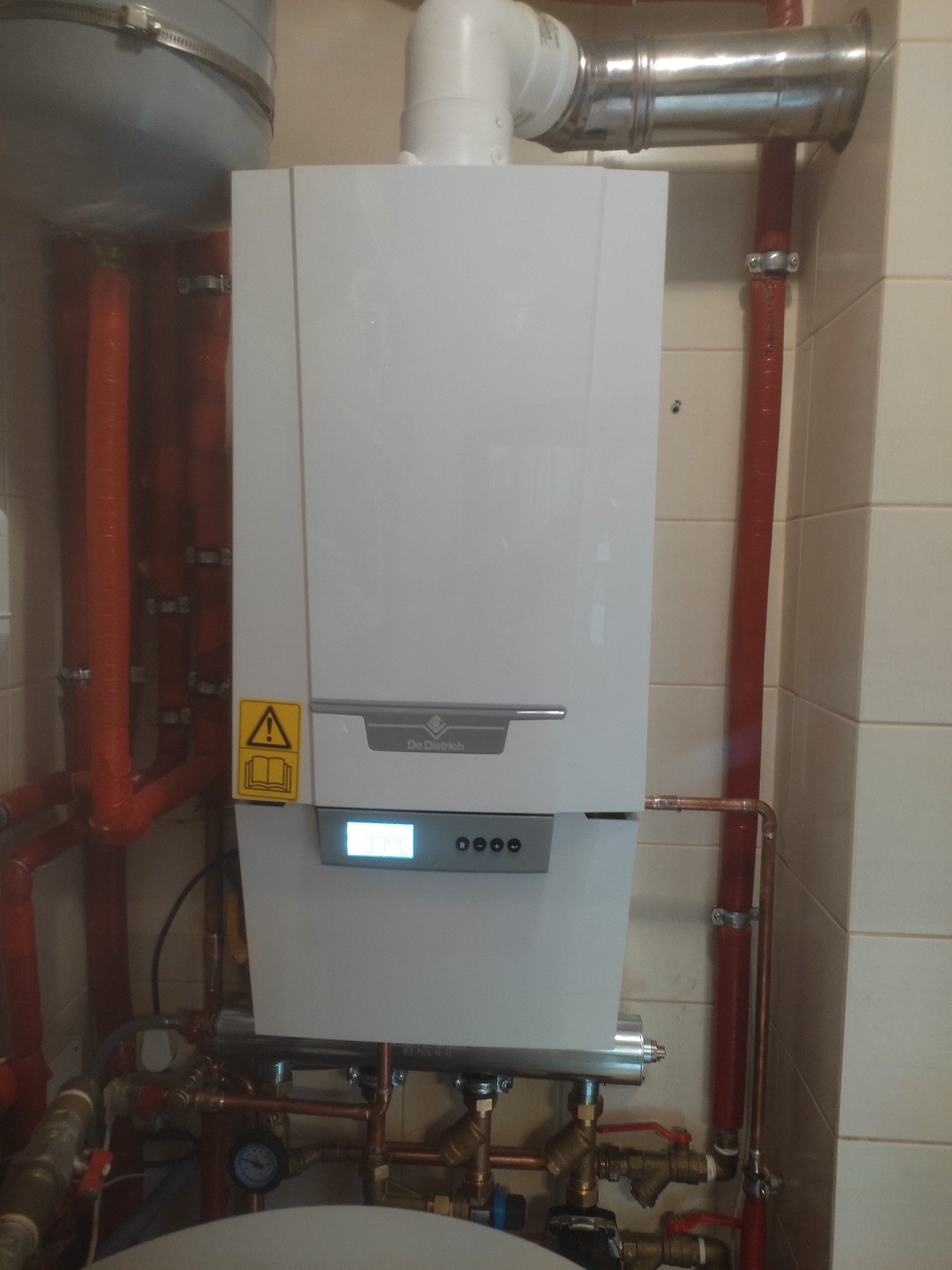 Biały piec gazowy marki De Dietrich zamontowany na ścianie z widocznymi rurami miedzianymi i plastikowymi, z żółtą naklejką ostrzegawczą po lewej stronie.