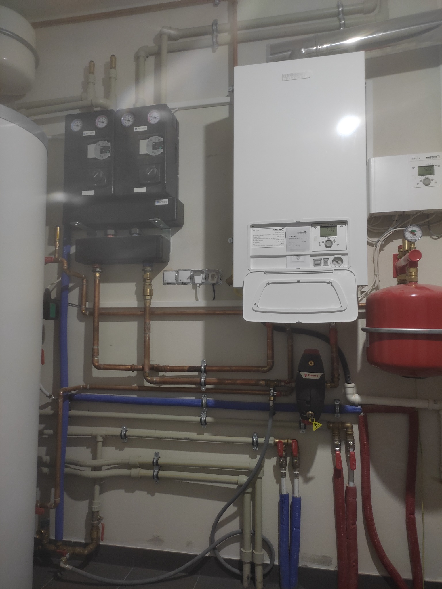 Biały piec gazowy z otwartą klapką na ścianie, obok czerwony zbiornik wyrównawczy, widoczne liczne rury miedziane i plastikowe, oraz czarne moduły sterujące z manometrami.