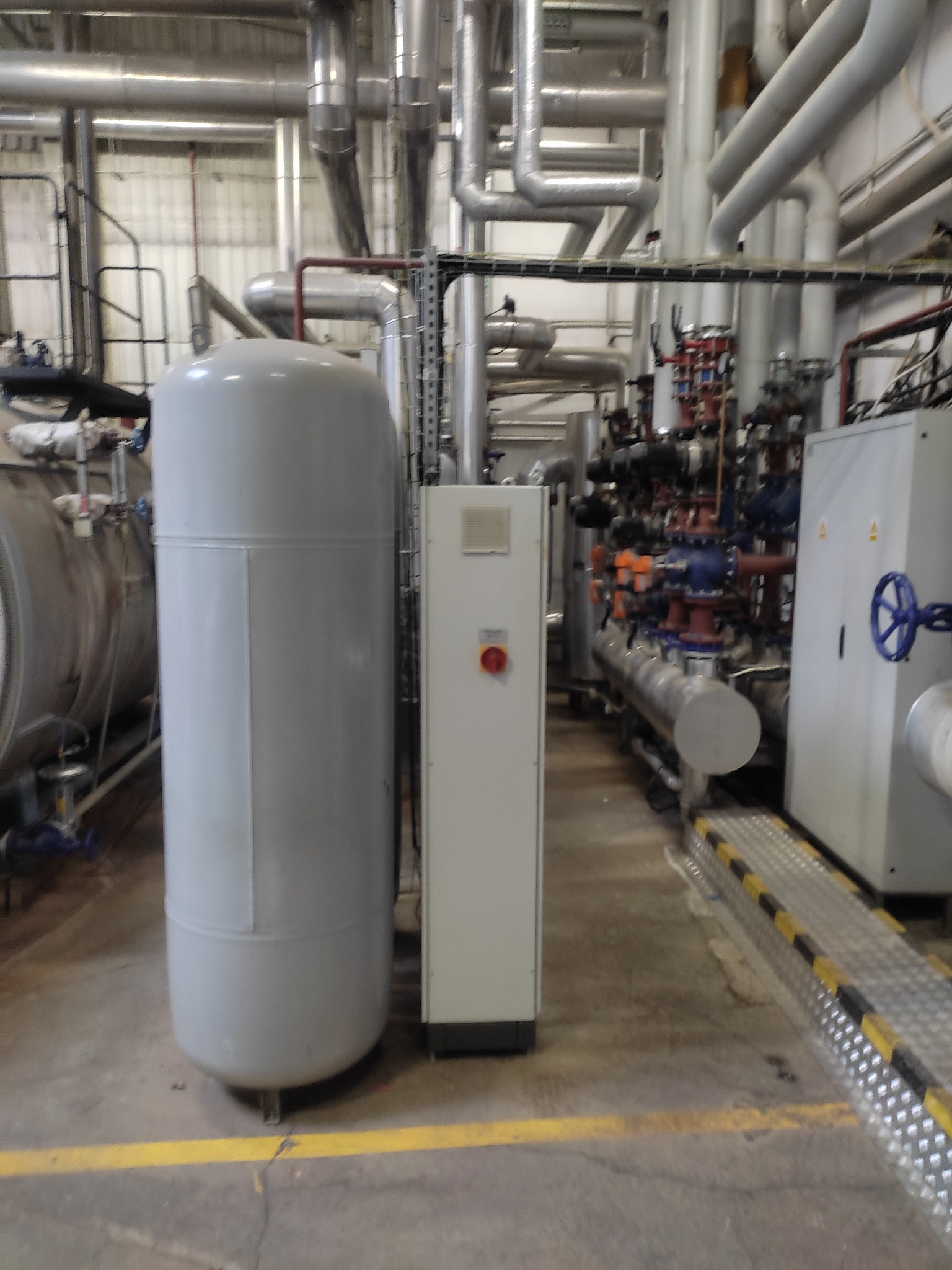 Instalacja przemysłowa z zbiornikiem ciśnieniowym, rurami, zaworami i szafą sterowniczą. Widoczne liczne rury i armatura w tle.