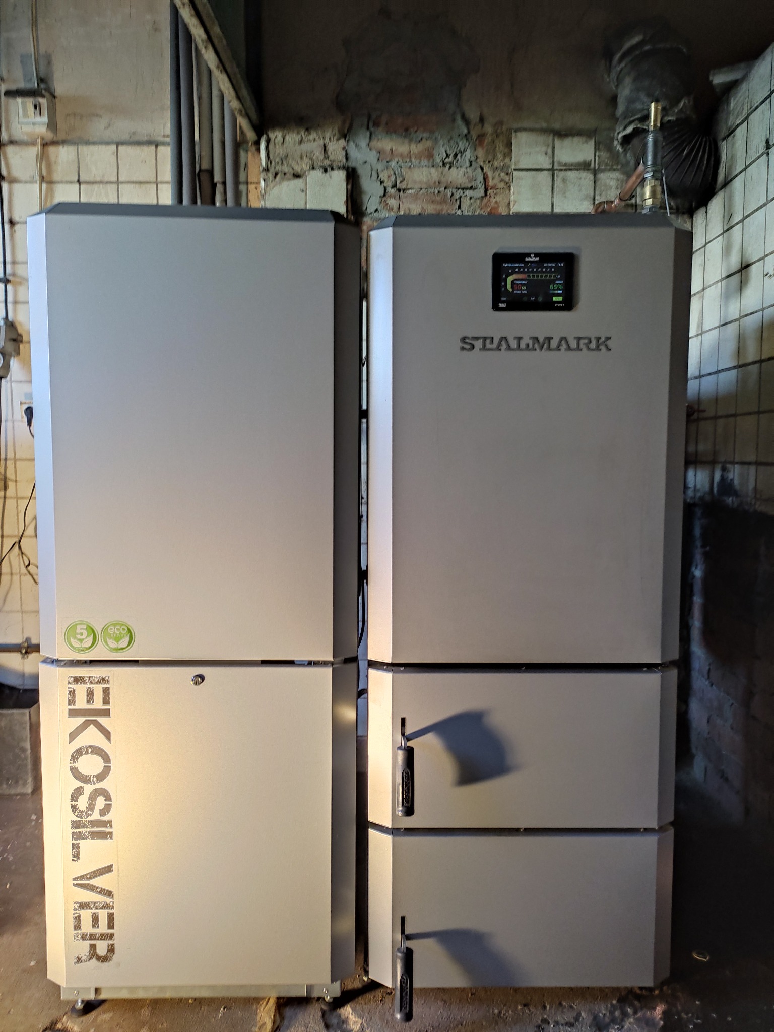 Dwa piece CO marki Stalmark EkoSilver, zainstalowane w kotłowni z widocznymi rurami i fragmentem instalacji wentylacyjnej.