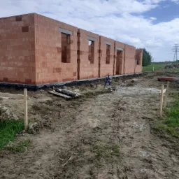 Wznoszone ściany domu z czerwonej cegły ceramicznej z widocznymi otworami okiennymi, zabezpieczone czarną folią izolacyjną na fundamencie, z widocznymi elementami instalacji i drewnianymi stemplami...