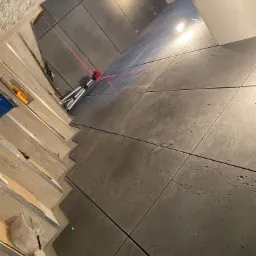 Świeżo ułożone, duże, szare płytki podłogowe z widoczną teksturą, wyznaczane laserowo, z elementami narzędzi i materiałów montażowych w tle.