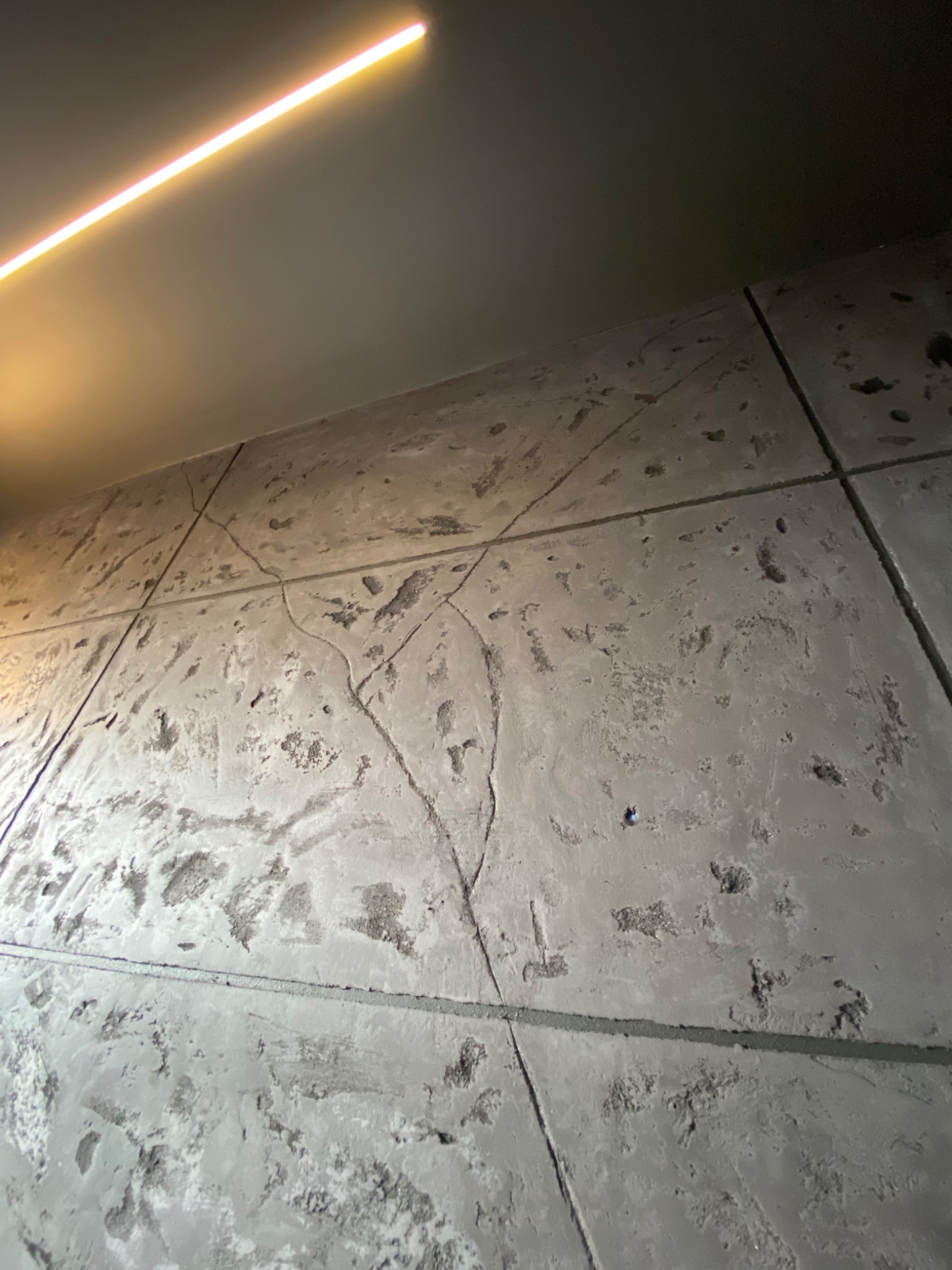 Ściana wykończona szarym tynkiem dekoracyjnym imitującym beton, podzielona na prostokątne segmenty z widocznymi fugami, oświetlona górnym, liniowym źródłem światła.