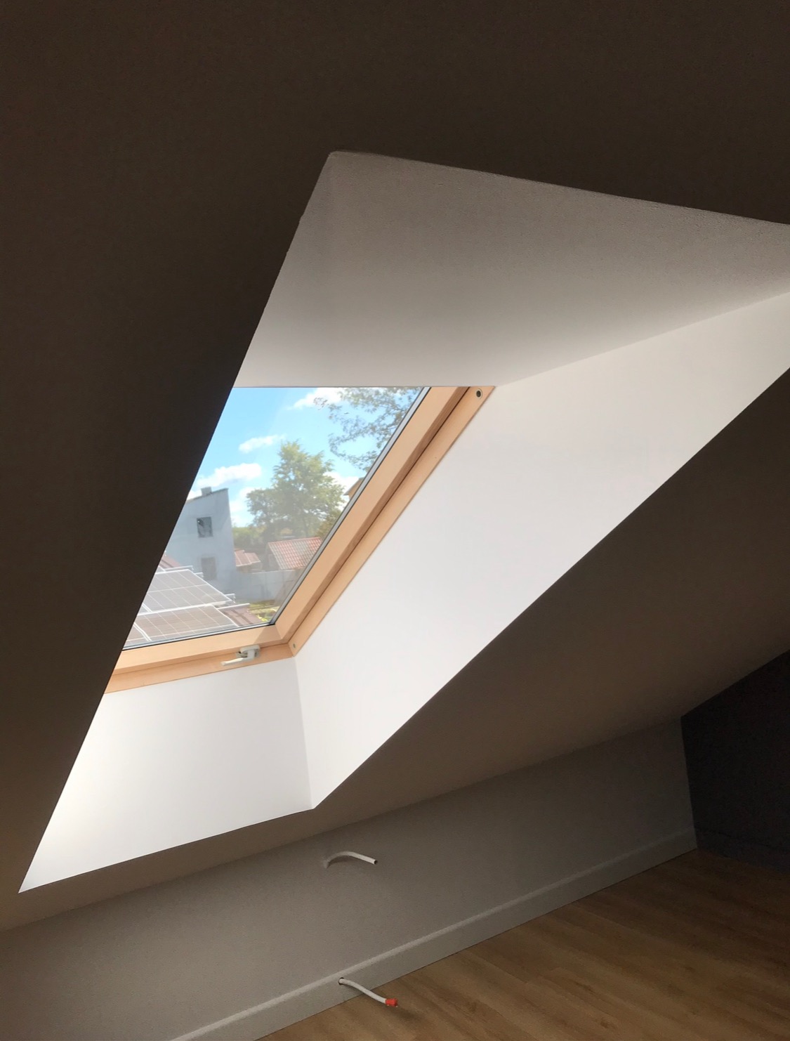 Poddasze z oknem dachowym w drewnianej ramie, widok na błękitne niebo i fragmenty zabudowań z panelami słonecznymi, widoczne kable wystające ze ściany, świeżo pomalowane ściany w odcieniach bieli...