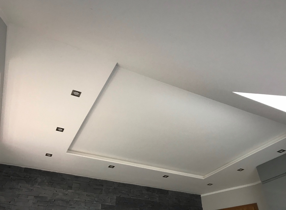 Biały sufit podwieszany z wpuszczonymi kwadratowymi lampkami halogenowymi, dekoracyjna ściana z szarej imitacji kamienia w dolnej części kadru.