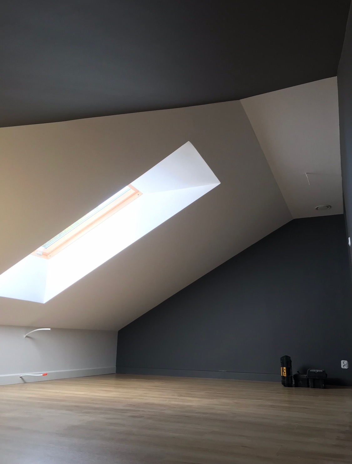 Poddasze po remoncie: świeżo pomalowane ściany w odcieniach szarości i bieli, drewnopodobna podłoga, okno dachowe wpuszczające jasne światło, narzędzia w rogu pomieszczenia.