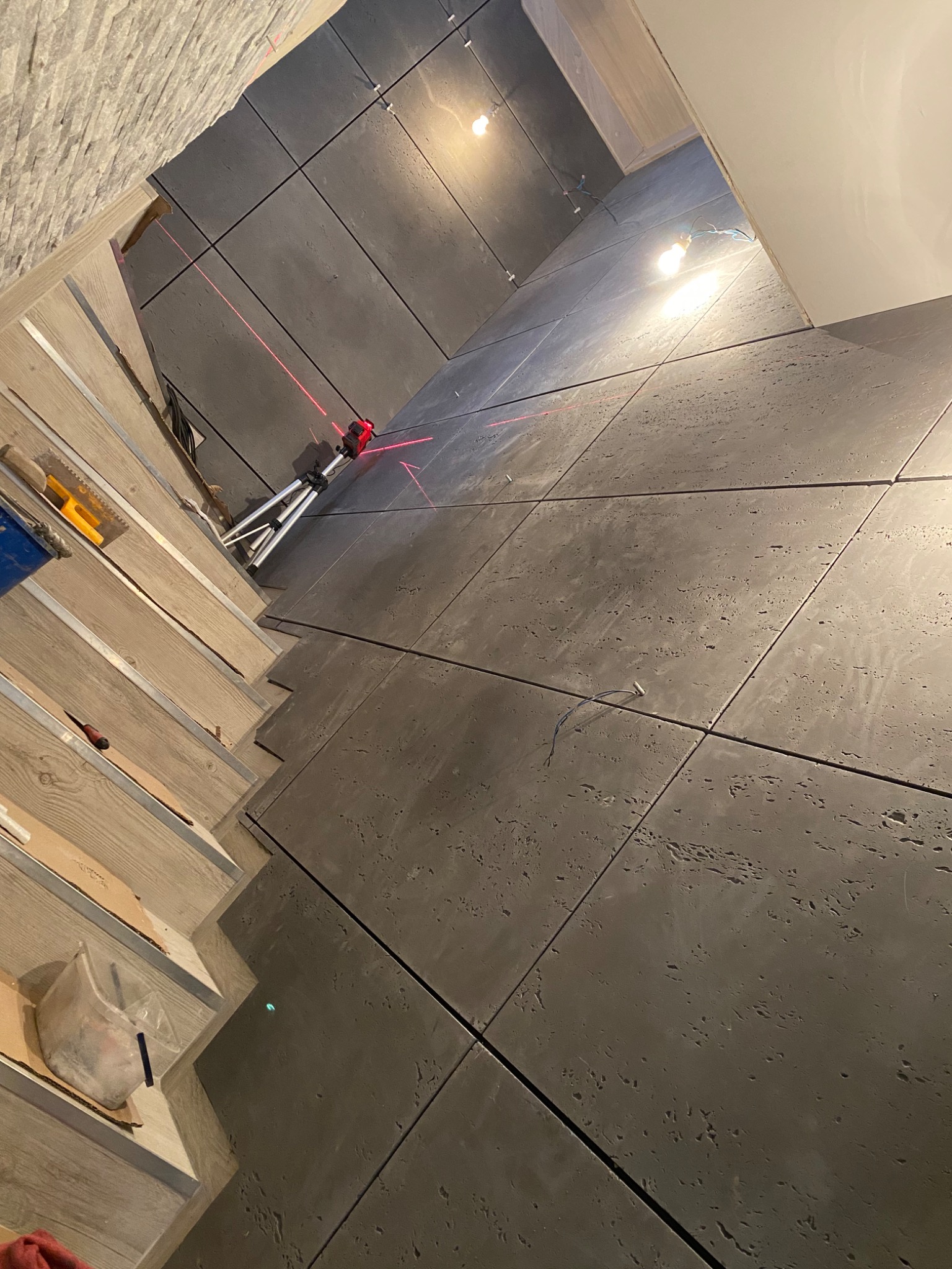 Świeżo ułożone, duże, szare płytki podłogowe z widoczną teksturą, wyznaczane laserowo, z elementami narzędzi i materiałów montażowych w tle.