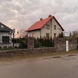 Murowane ogrodzenie z elementami kutymi przed dwoma domami, widok z ulicy w pochmurny dzień.