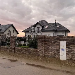 Murowane ogrodzenie z kutymi elementami przed dwoma domami jednorodzinnymi o nowoczesnej architekturze, sfotografowane o zmierzchu.