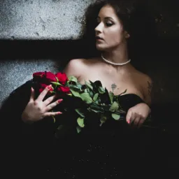 Kobieta w czarnej sukni z perłowym naszyjnikiem, leżąca na schodach z bukietem czerwonych róż, z tatuażem róży na dekolcie, spoglądająca w lewo.