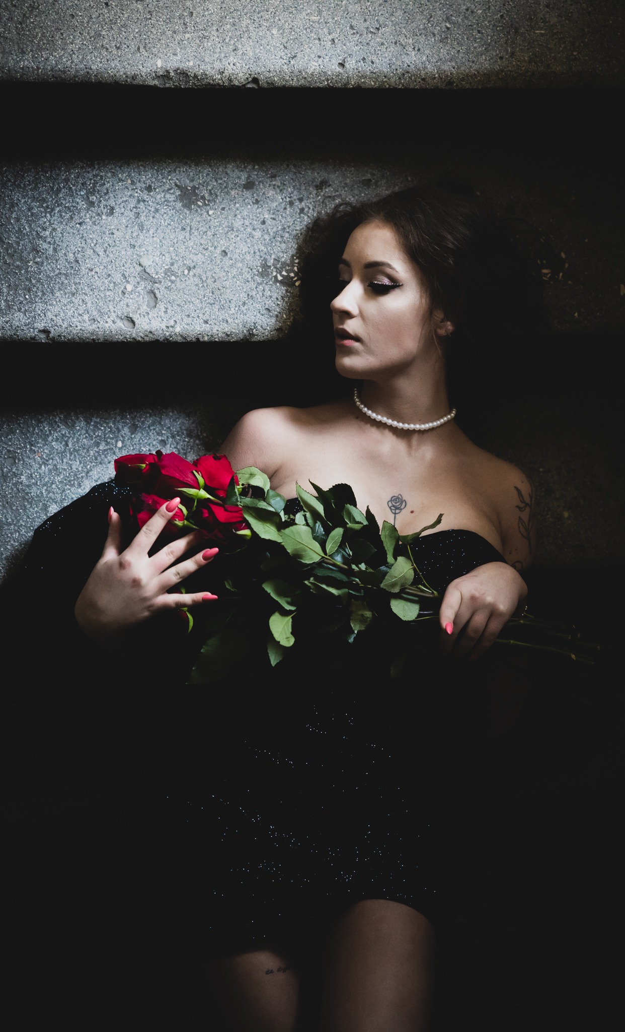 Kobieta w czarnej sukni z perłowym naszyjnikiem, leżąca na schodach z bukietem czerwonych róż, z tatuażem róży na dekolcie, spoglądająca w lewo.