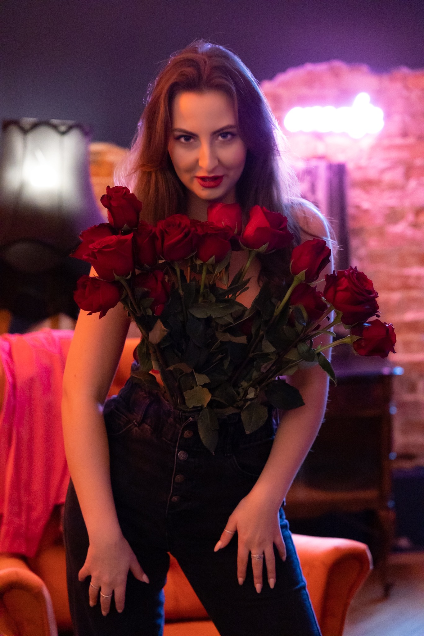 Kobieta trzyma bukiet czerwonych róż, ubrana w czarne jeansy na tle wnętrza z neonowym światłem i lampą.
